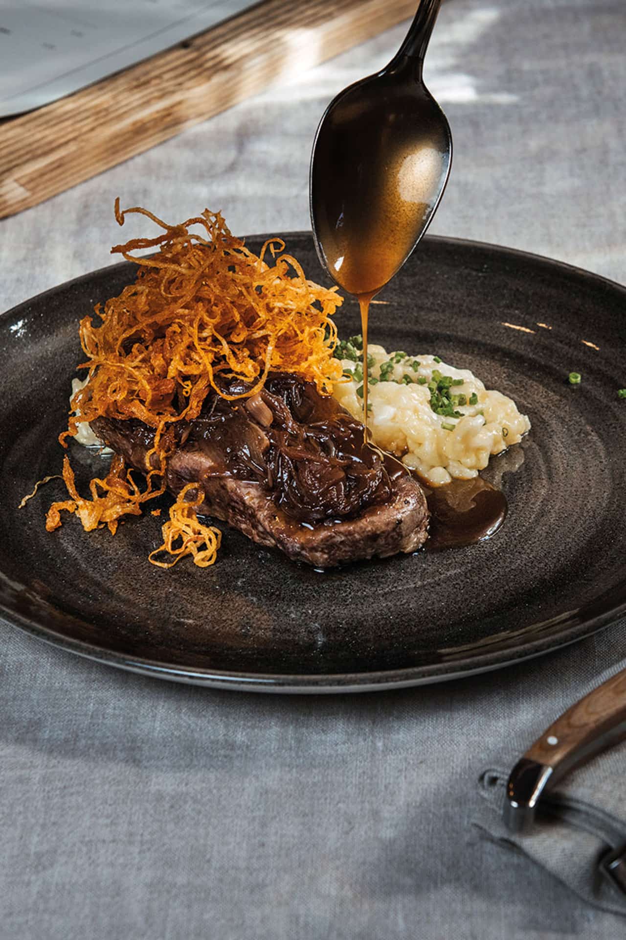Detailaufnahme eines Zwiebelrostbratens mit Kässpatzen und einem Saucenlöffel