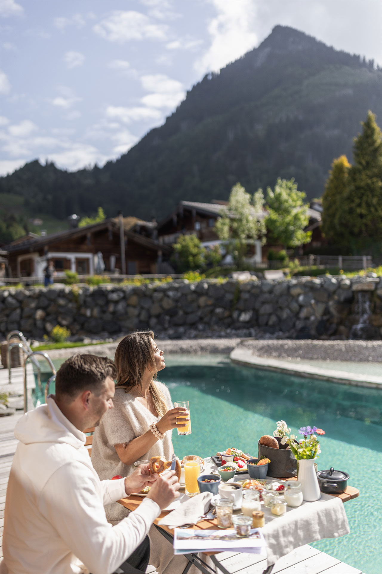 Ein Paar genießt ein Frühstück im Freien am Rand eines Schwimmbeckens, umgeben von Bergen. Auf dem Tisch sind verschiedene Speisen und Getränke, darunter Obst und Brötchen, dekoriert mit Blumen. Die Atmosphäre ist entspannt und sonnig.