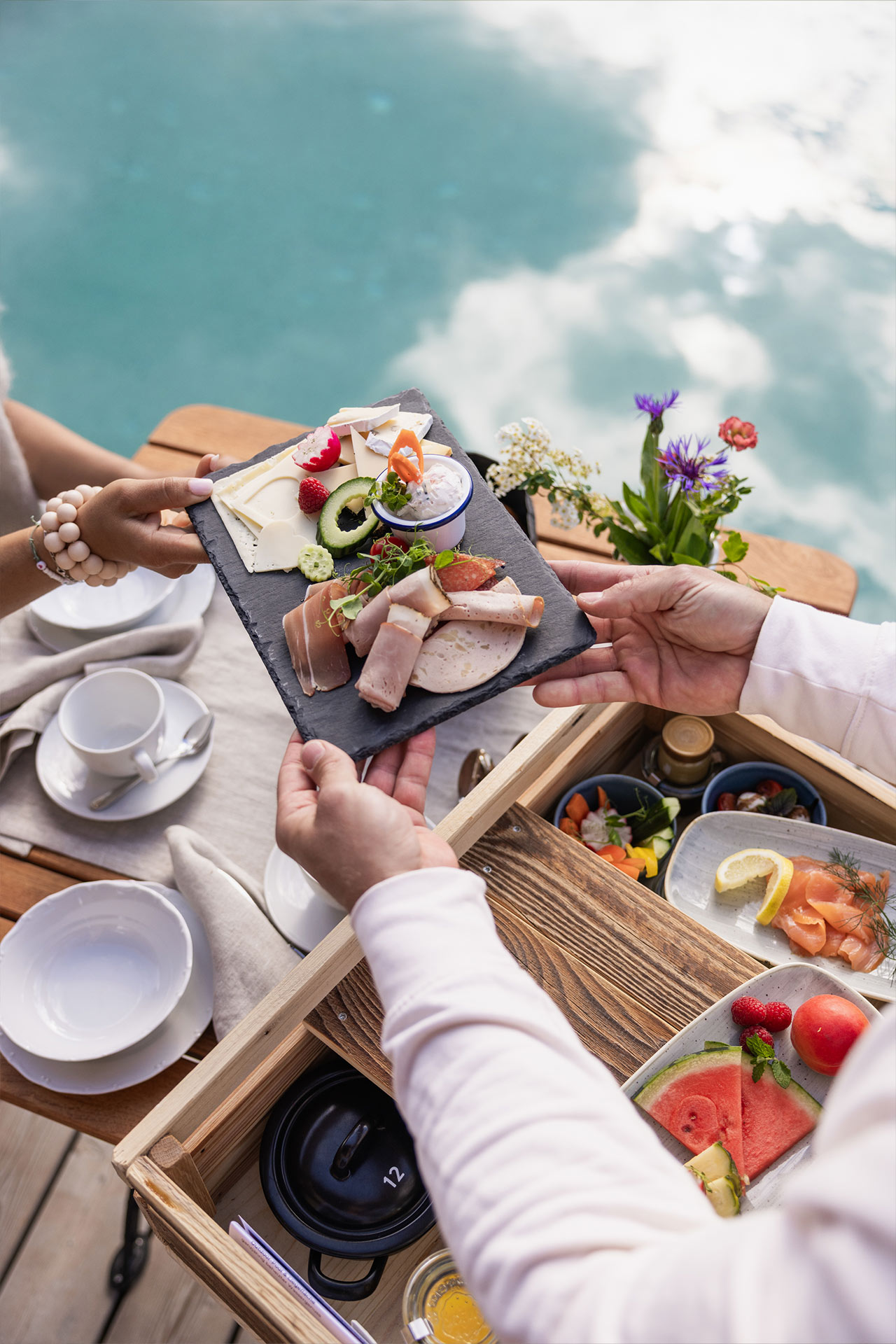Eine Person reicht einen schwarzen Teller mit Aufschnitt, Käse und Gemüse an eine andere Person. Im Hintergrund sind Blumen und eine Tasse Kaffee auf einem Tisch zu sehen. Der Tisch steht am Wasser.