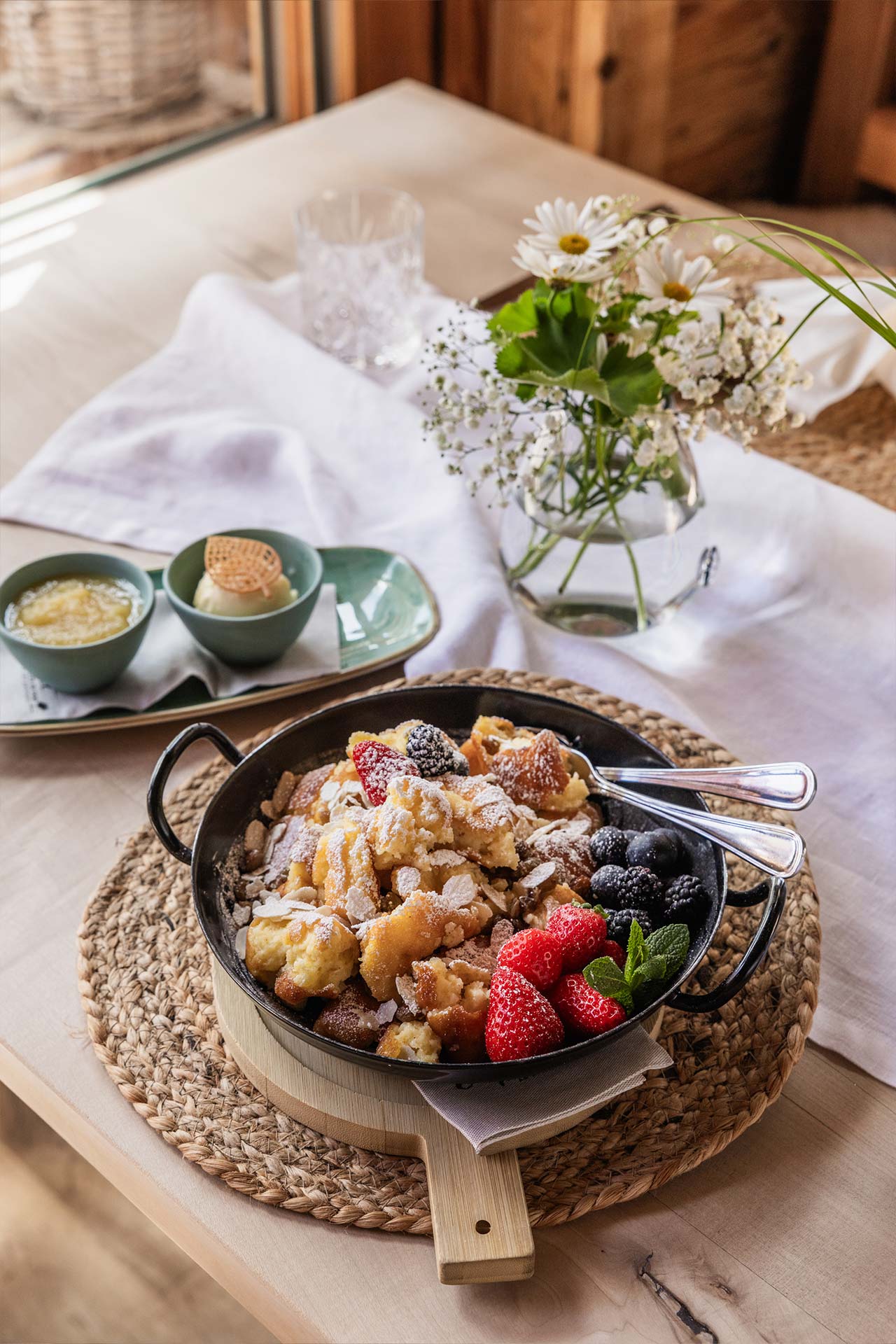 Eine Schüssel mit einem süßen Gericht, garniert mit frischen Beeren wie Erdbeeren und schwarzen Johannisbeeren, liegt auf einem rustikalen Tisch. Neben der Schüssel stehen zwei kleine Teller mit Desserts und einer Blumenvase mit weißen Blumen.