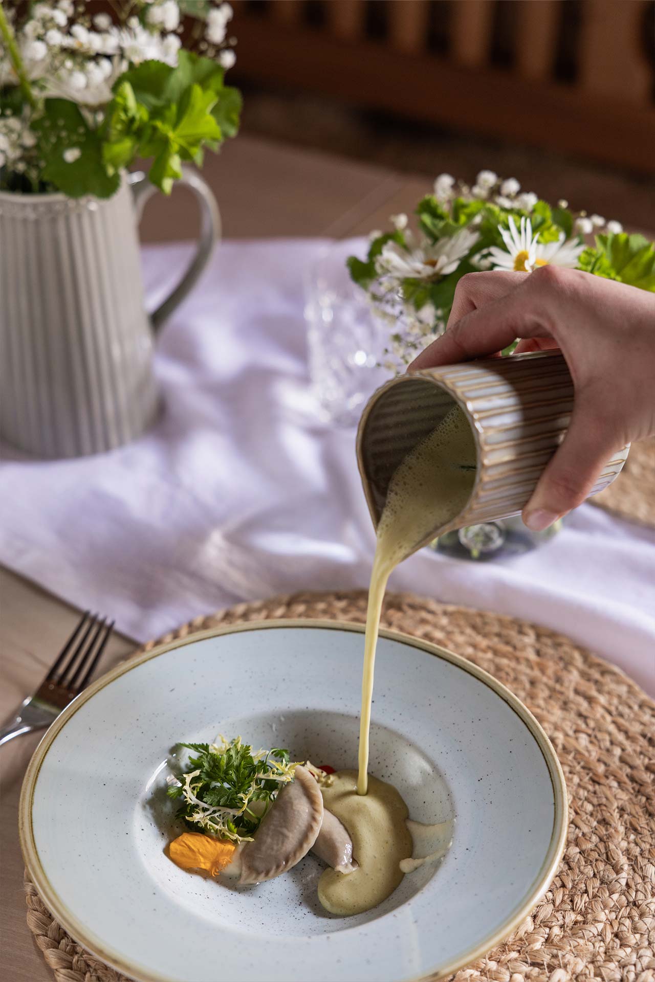 Eine Hand gießt eine cremige Sauce aus einem Becher auf einen eleganten Teller mit einer kunstvoll angerichteten Speise. Der Teller enthält eine kleine Portion Gemüse und Kräuter. Im Hintergrund sind Blumen in einer Vase und eine schön gedeckte Tischdekoration zu sehen.