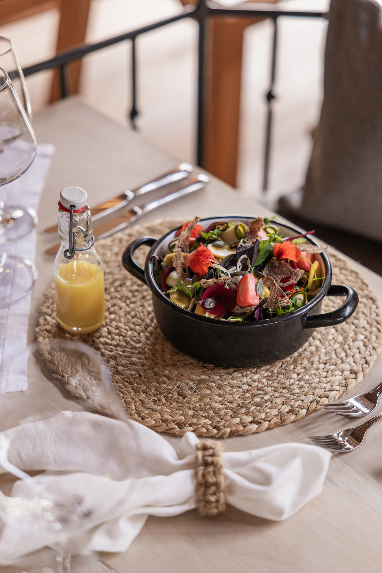 Ein appetitlich angerichteter Salat in einem schwarzen Topf steht auf einem runden, geflochtenen Platzset. Neben dem Topf befindet sich eine kleine Flasche mit gelber Sauce. Der Tisch ist elegant gedeckt mit Besteck und einem Serviettenring.