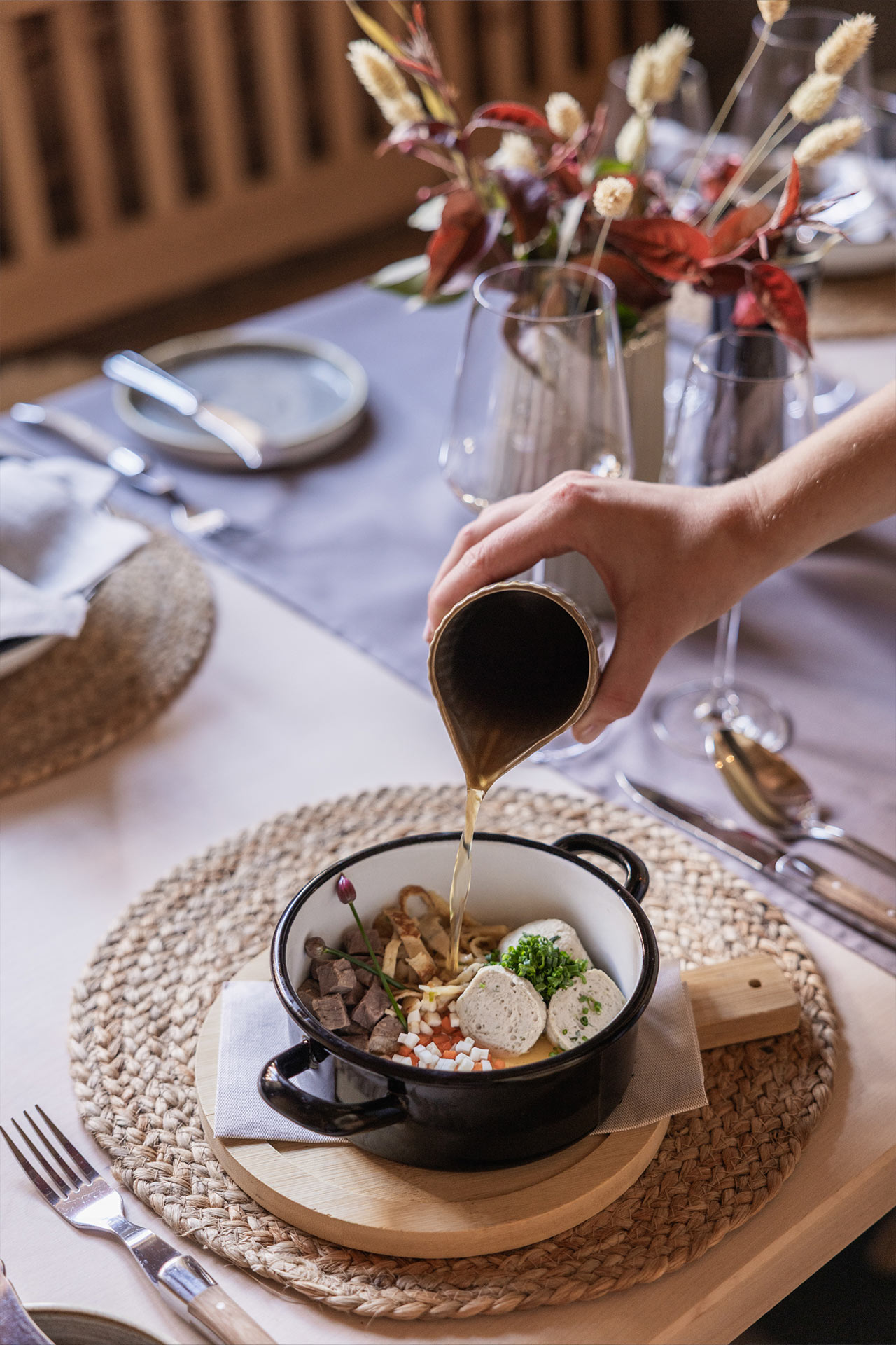 Eine Hand gießt Brühe aus einem Kännchen in eine schwarze Schüssel mit verschiedenen Zutaten, darunter Fleisch, Gemüse und Kräuter. Der Tisch ist elegant gedeckt mit Besteck, Gläsern und Dekoration aus Trockenblumen.
