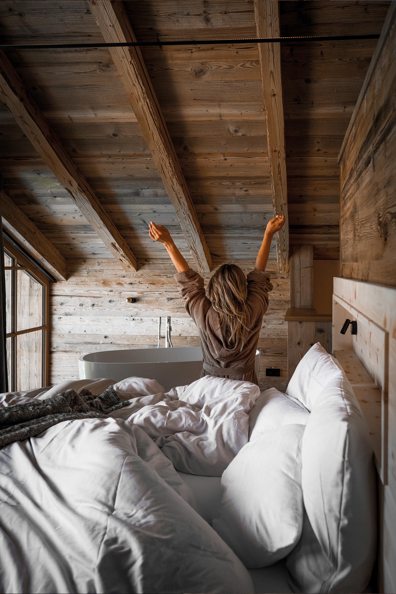 eine junge Frau streckt sich nach dem Aufwachen im Bett ihres Chalets. 