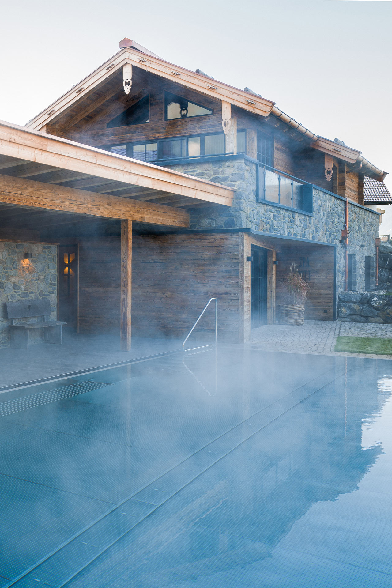 Ein modernes Haus aus Stein und Holz steht neben einem Dampfbad, das von feinem Nebel umgeben ist. Das Gebäude hat große Fenster und ein Stufendesign, während der Pool ruhig im Vordergrund liegt. Der gesamte Bereich strahlt eine entspannende Atmosphäre aus.