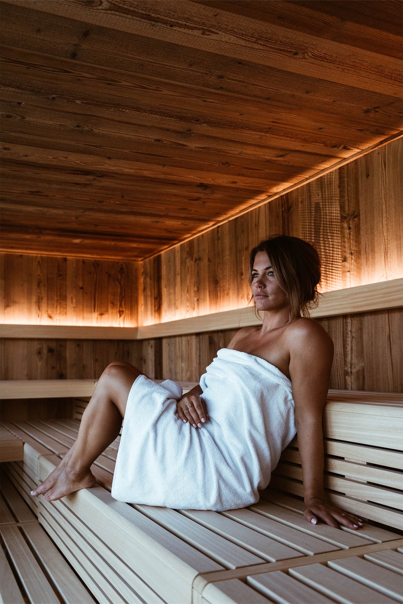 Eine Frau sitzt entspannt in einer Holzsauna, umhüllt von einem weißen Handtuch. Sie lehnt sich leicht zurück, während warmes Licht die Wände sanft beleuchtet. Der Raum vermittelt eine ruhige und gemütliche Atmosphäre, ideal zum Entspannen.