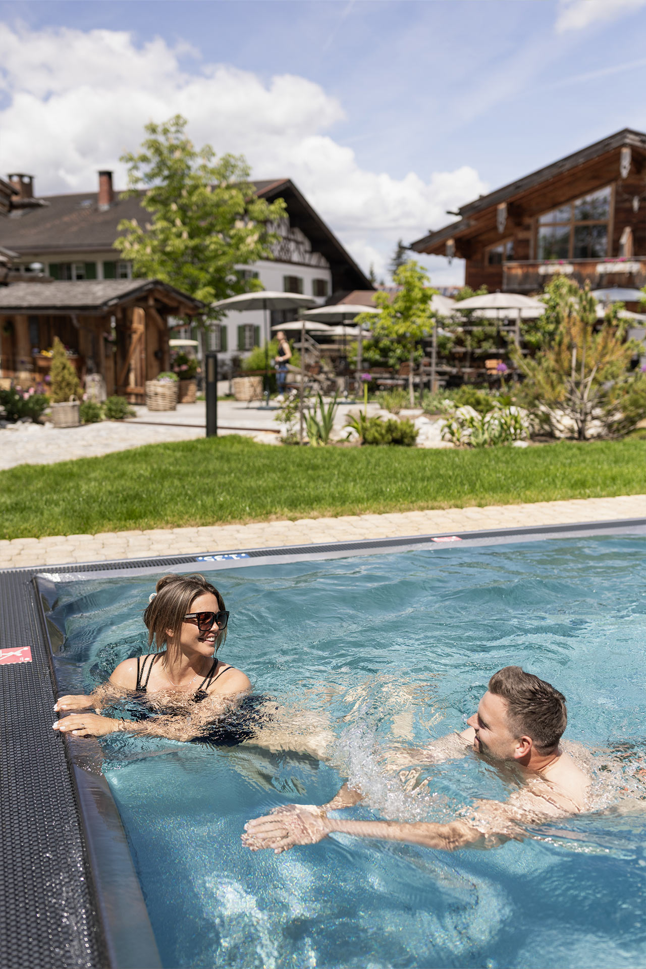 Ein Paar entspannt im Pool an einem sonnigen Tag. Im Hintergrund sind ein Garten und ein Gebäude mit Holzelementen zu sehen. Der Himmel ist blau mit einigen Wolken.