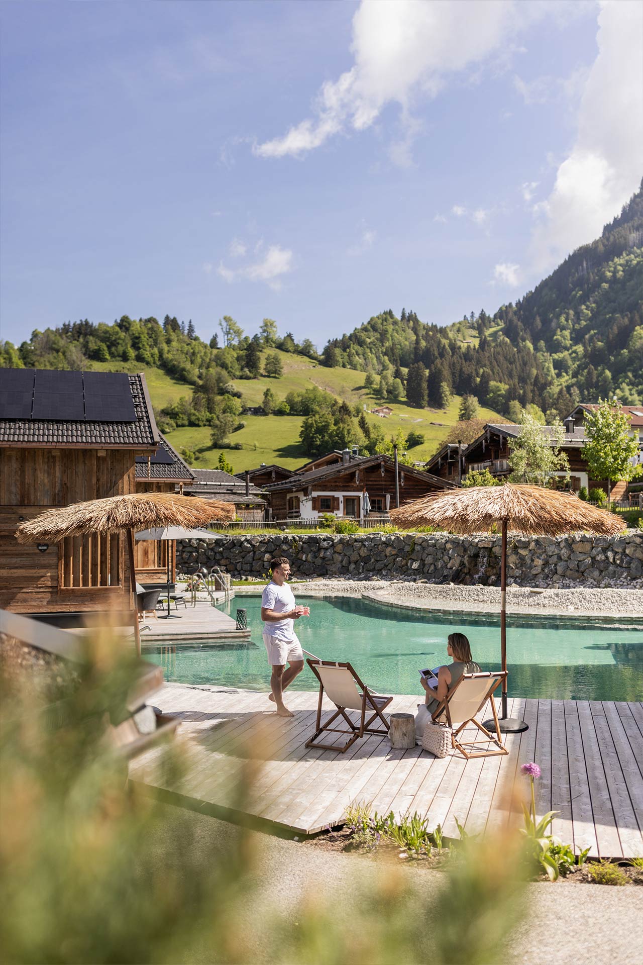 Ein Mann und eine Frau sitzen am Rand eines Pools, umgeben von Hütten und grünen Bergen. Es ist sonnig, und beide entspannen sich unter Sonnenschirmen. Die Atmosphäre ist ruhig und idyllisch.