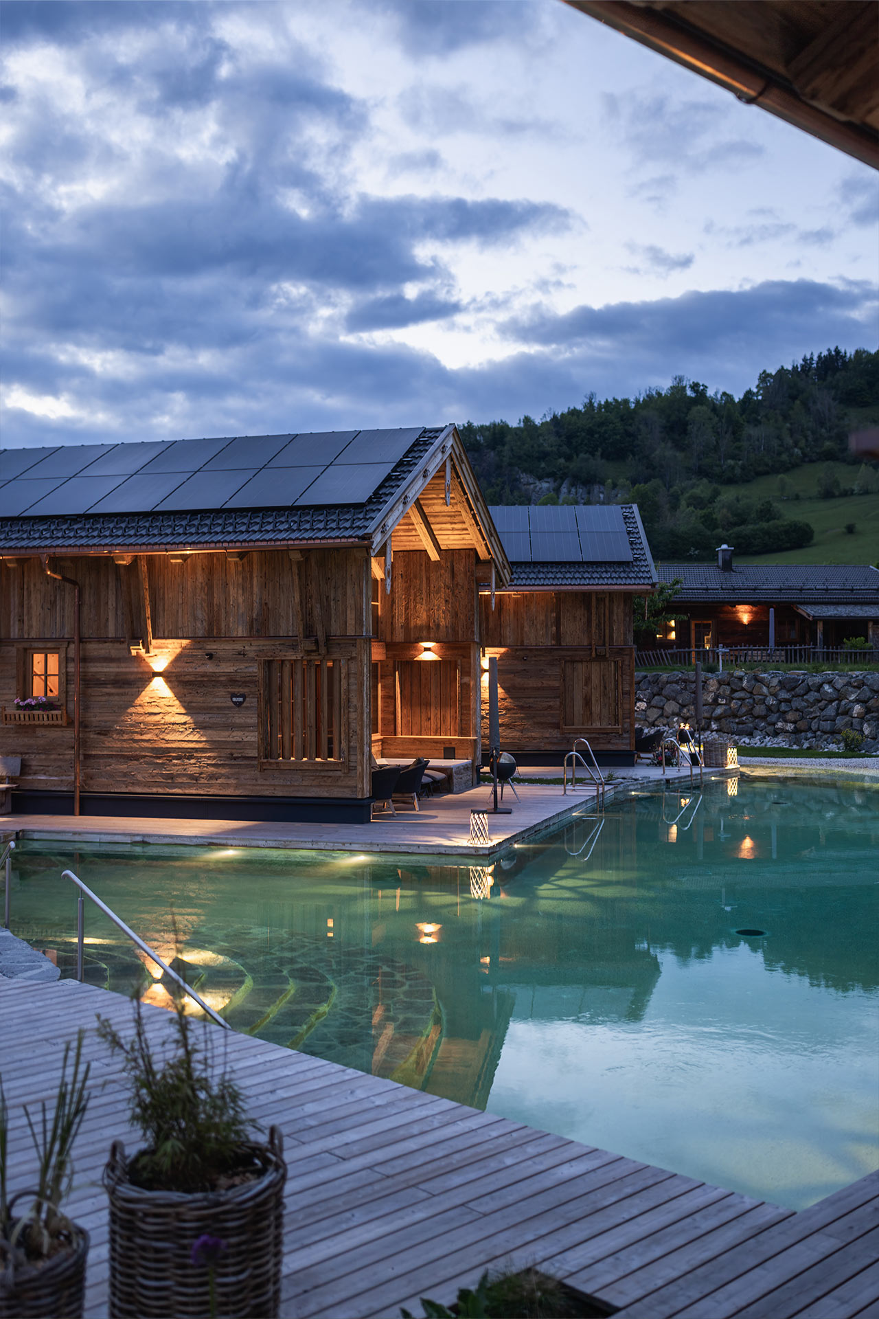 Ein Holzgebäude mit Solarpanelen steht am Ufer eines beleuchteten Schwimmbeckens. Im Hintergrund sind sanfte Hügel und ein bewölkter Himmel zu sehen, während die Dämmerung einsetzt. Die Umgebung wirkt ruhig und einladend.