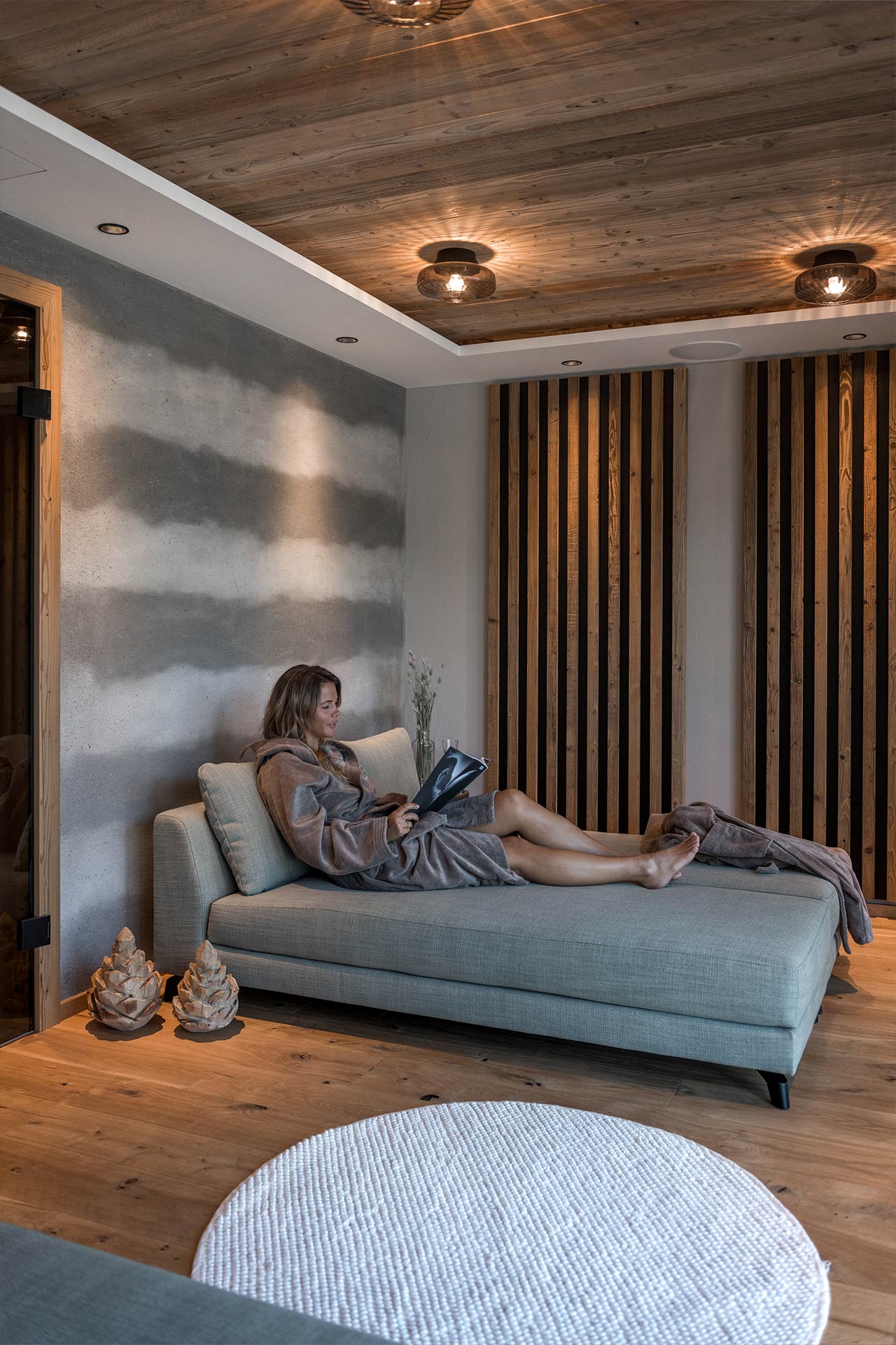 Eine entspannte Frau sitzt auf einer Couch in einem modernen Raum mit Holzdetails. Sie trägt einen Bademantel und liest in einem Buch. Der Raum hat gedämpftes Licht und eine gemütliche Atmosphäre, ideal zum Entspannen.