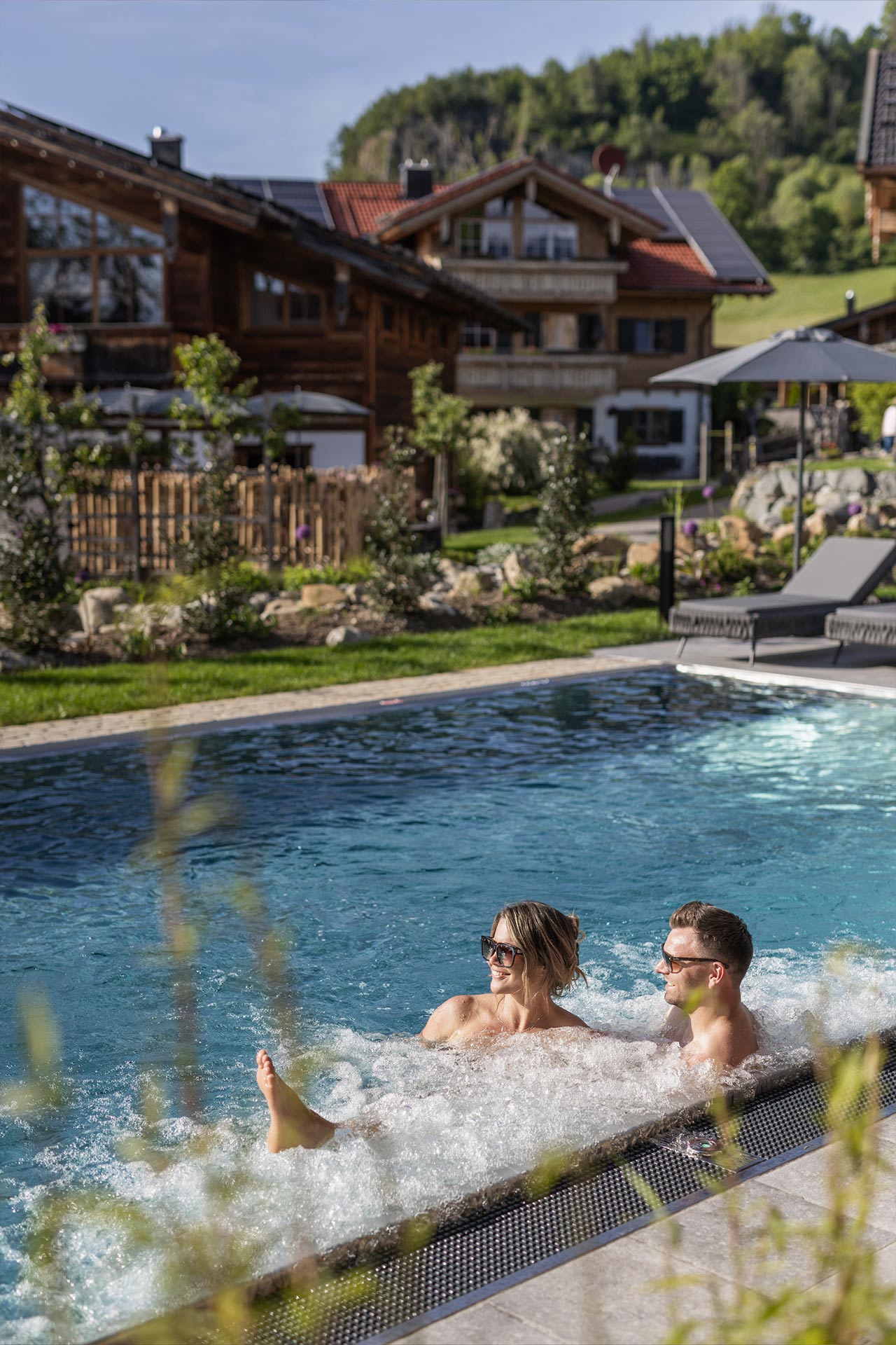 Ein Paar sitzt entspannt am Ende eines Schwimmbeckens mit sprudelndem Wasser. Im Hintergrund sind Holzhäuser und grüne Hügel zu sehen. Es ist ein sonniger Tag, und die Umgebung ist mit Pflanzen und Sonnenschirmen dekoriert.
