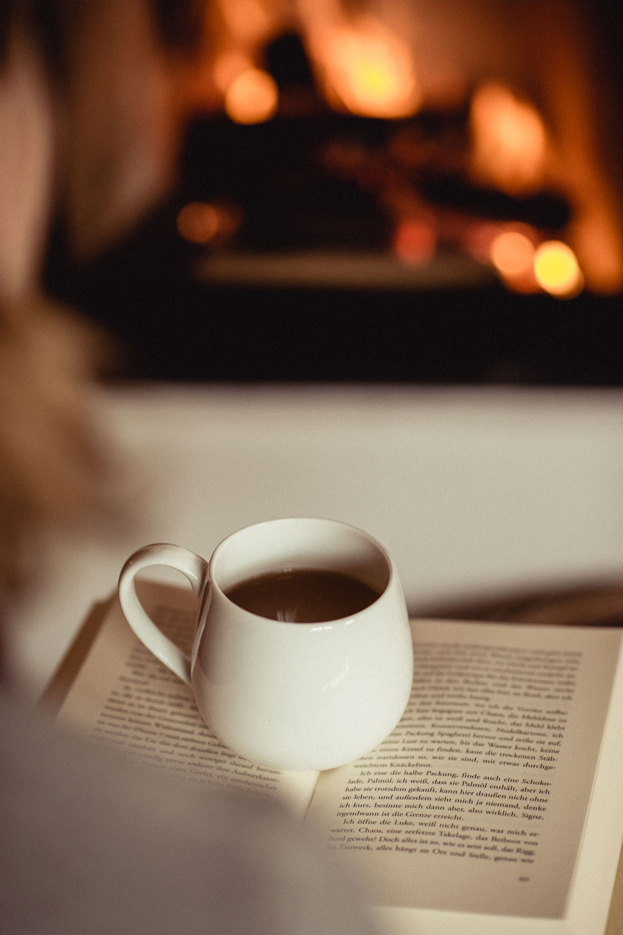 Detailaufnahme einer Teetasse und einem Buch vor einem brennenden Kaminofen