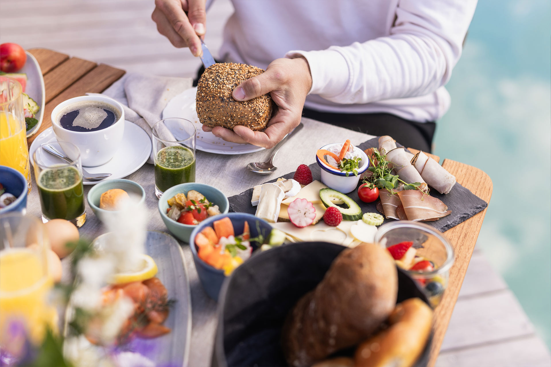 Eine Person schneidet ein Vollkornbrötchen. Vor ihnen steht ein reichhaltiges Frühstücksbuffet mit diversen Speisen wie Obst, Gemüse, Aufschnitt und Joghurt sowie Getränken wie Kaffee und Säften. Der Tisch ist liebevoll gedeckt.