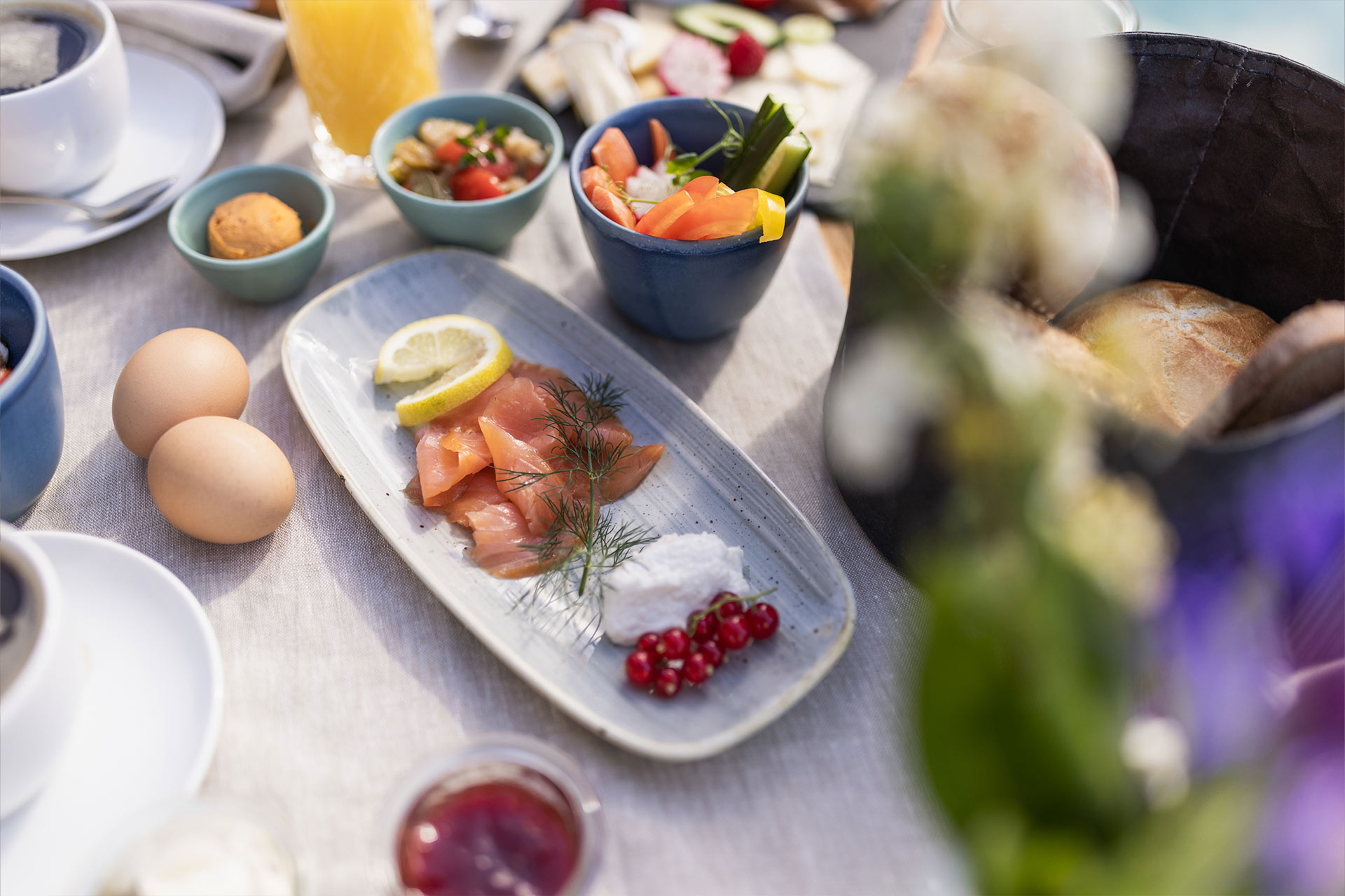 Ein gedeckter Frühstückstisch mit verschiedenen Speisen. Im Vordergrund liegt ein Teller mit Räucherlachs, Zitrone, Frischkäse und roten Johannisbeeren. Daneben befinden sich Eier, kleine Gerichte in Schalen und ein Korb mit Brötchen. Das Gesamtbild vermittelt eine einladende Frühstücksatmosphäre.