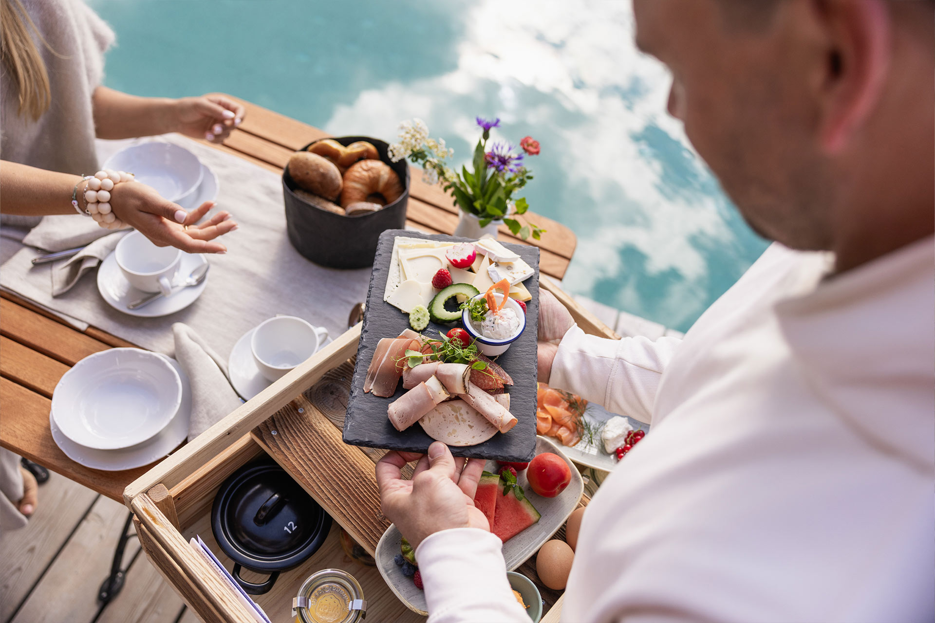 Ein Mann hält ein Tablett mit verschiedenen Speisen, darunter Aufschnitt und Obst. Im Hintergrund ist ein Tisch mit Kaffeetassen und frischen Brötchen zu sehen, während eine Frau mit gestreckter Hand spricht. Der Ozean spiegelt den Himmel wider.