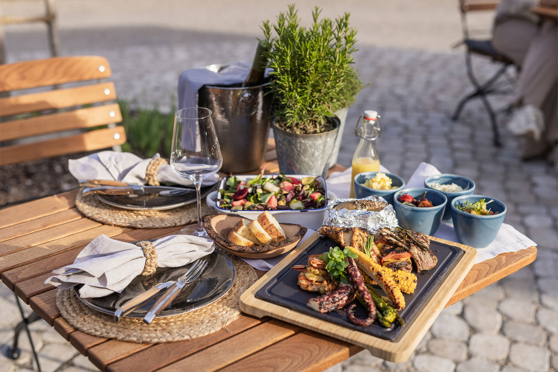 Ein schön gedeckter Tisch im Freien mit verschiedenen Speisen. Im Vordergrund liegt eine große Platte mit Grillfleisch und Gemüse. Daneben stehen Schalen mit Salat, Dips und Getränken. Eine Pflanze in einem Topf ergänzt die Tischdekoration.