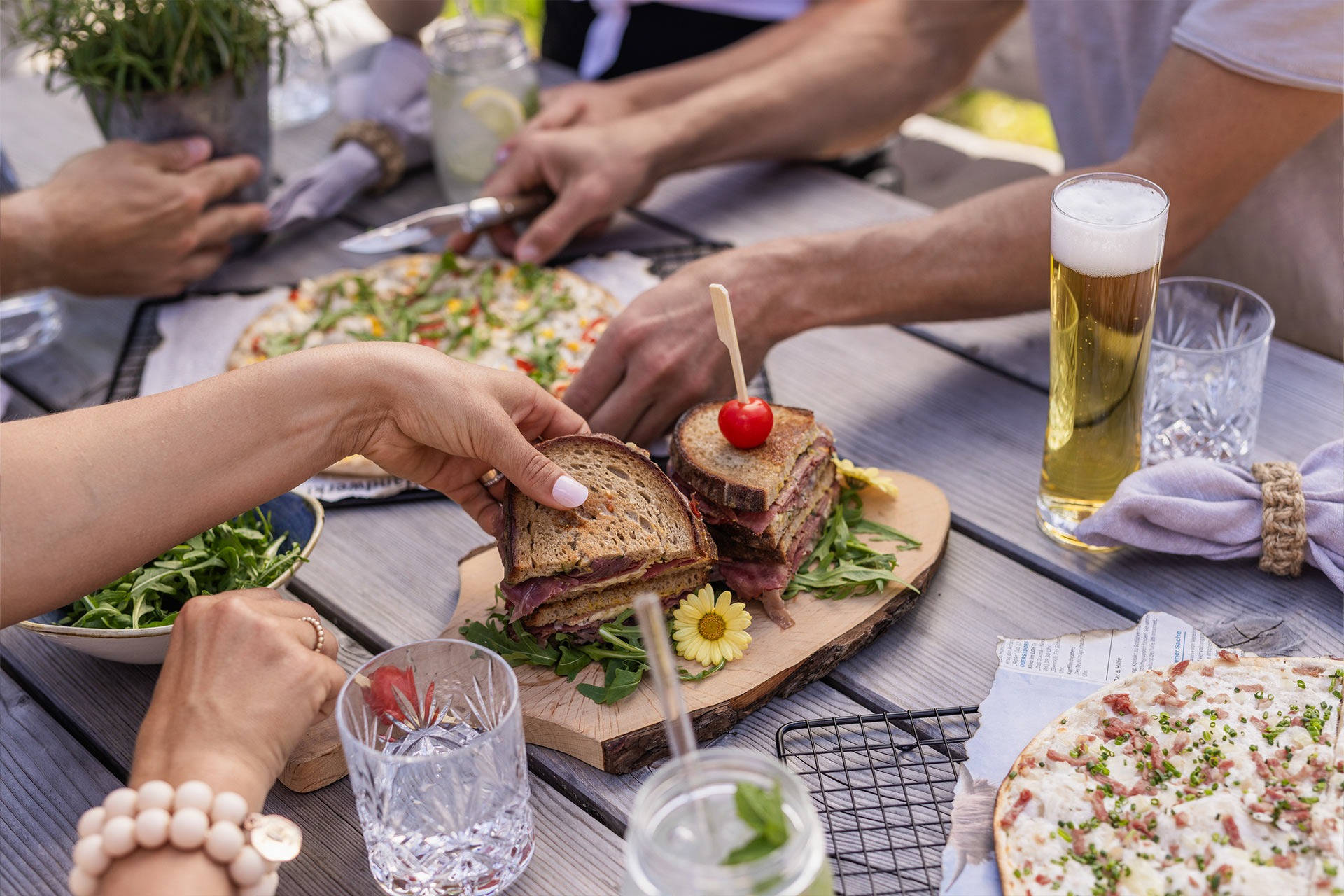 Eine Gruppe von Menschen sitzt an einem Tisch im Freien und genießt Essen. In der Mitte liegt ein Sandwich mit einem kleinen roten Schirmchen, umgeben von Salat, Getränken und einer Pizza. Einige Hände greifen nach dem Sandwich oder halten Gläser.