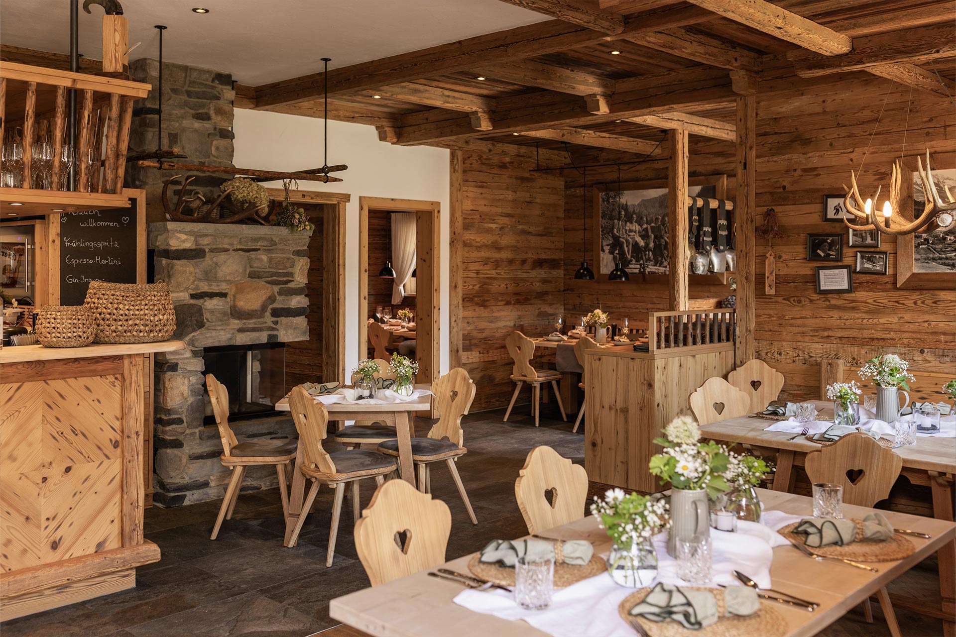 Ein einladendes Restaurant mit Holzinterieur, rustikalen Tischen und Stühlen. In der Ecke steht ein Kamin, und an den Wänden hängen Bilder. Tische sind mit Blumensträußen und Geschirr gedeckt, was eine gemütliche Atmosphäre schafft.