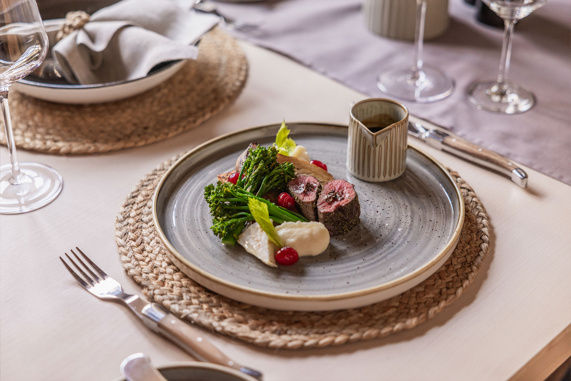 Ein eleganter Teller mit einem Gericht, das aus Rinderfilet, grünem Gemüse, kleinen roten Beeren und einer cremigen Beilage besteht. Der Teller ist auf einem rustikalen Platzdeckchen arrangiert, umgeben von Besteck und Weingläsern auf einem schön gedeckten Tisch.