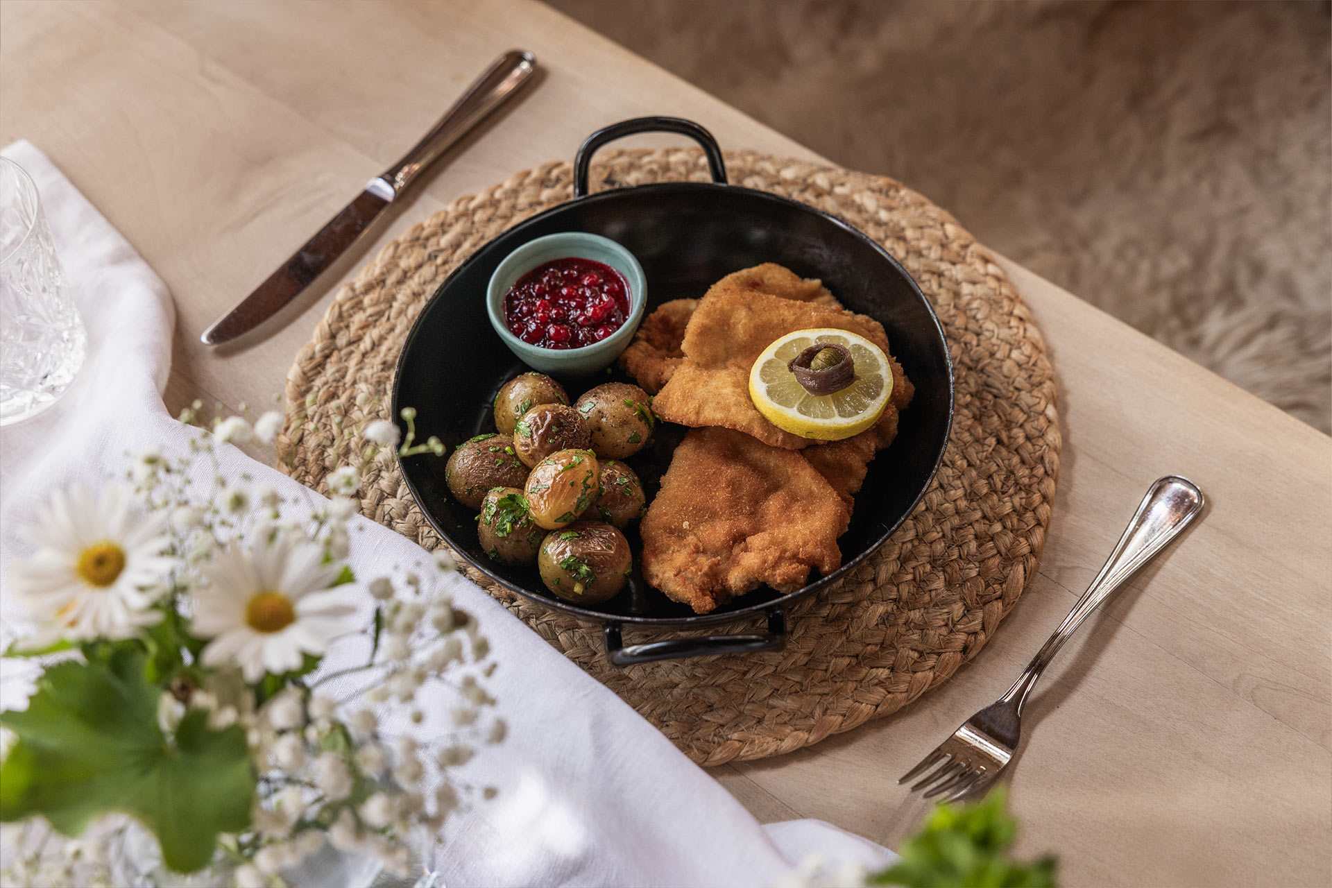 Ein schwarzes Serviergericht mit paniertem Fisch, einer Zitronenscheibe und kleinen, gebratenen Kartoffeln. Neben dem Gericht befindet sich eine Schale mit roter Soße. Der Teller steht auf einem runden Tischset aus Naturfasern.