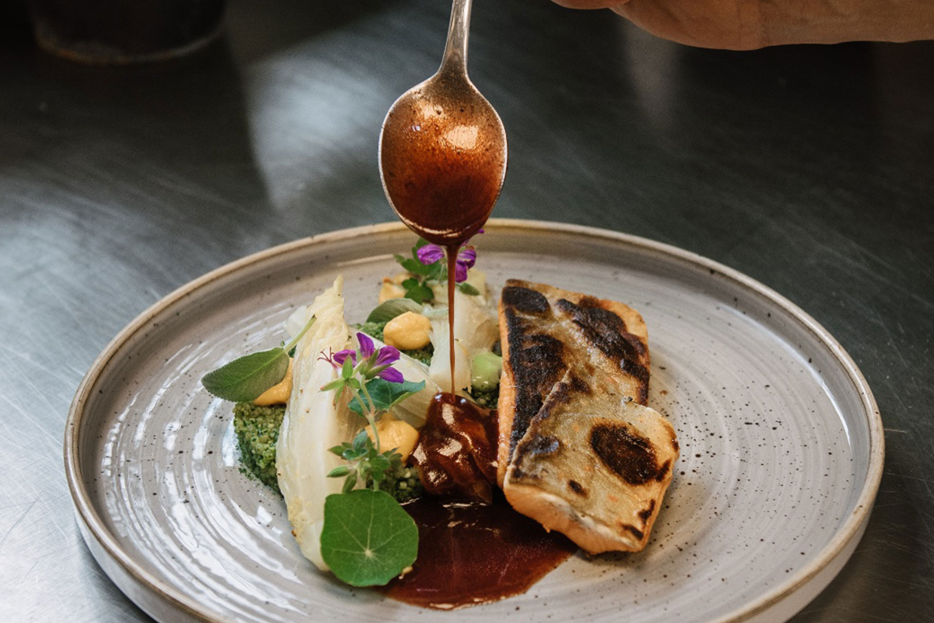 A hand pours sauce onto a beautifully plated dish featuring grilled fish, accompanied by vibrant vegetables, herbs, and edible flowers. The arrangement highlights a mix of textures and colors, emphasizing a gourmet presentation.