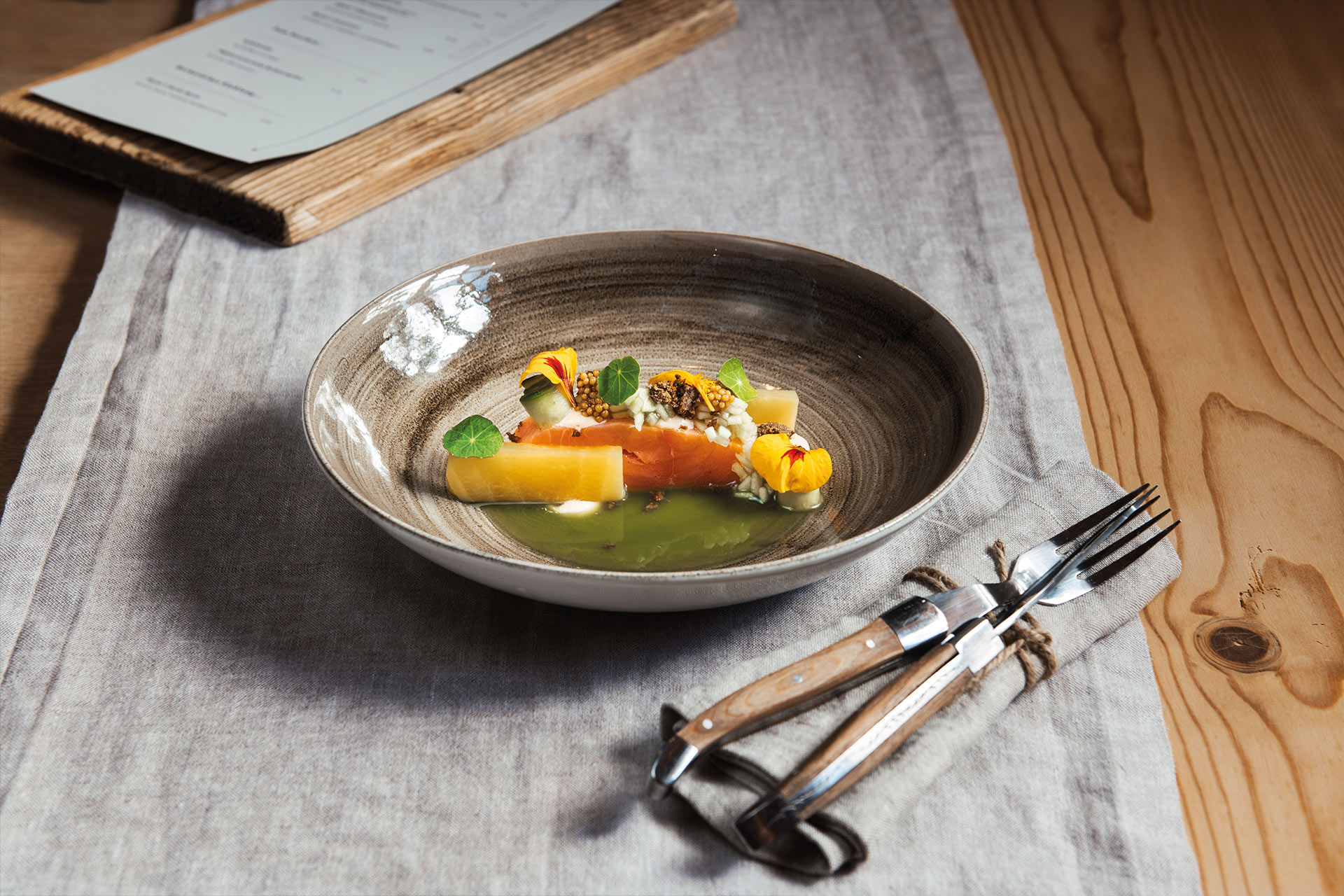 A beautifully arranged dish in a deep bowl, featuring colorful ingredients such as salmon, fresh herbs, and edible flowers, accompanied by a vibrant green sauce. The setting includes a cloth napkin and utensils beside the bowl, which rests on a textured table surface.