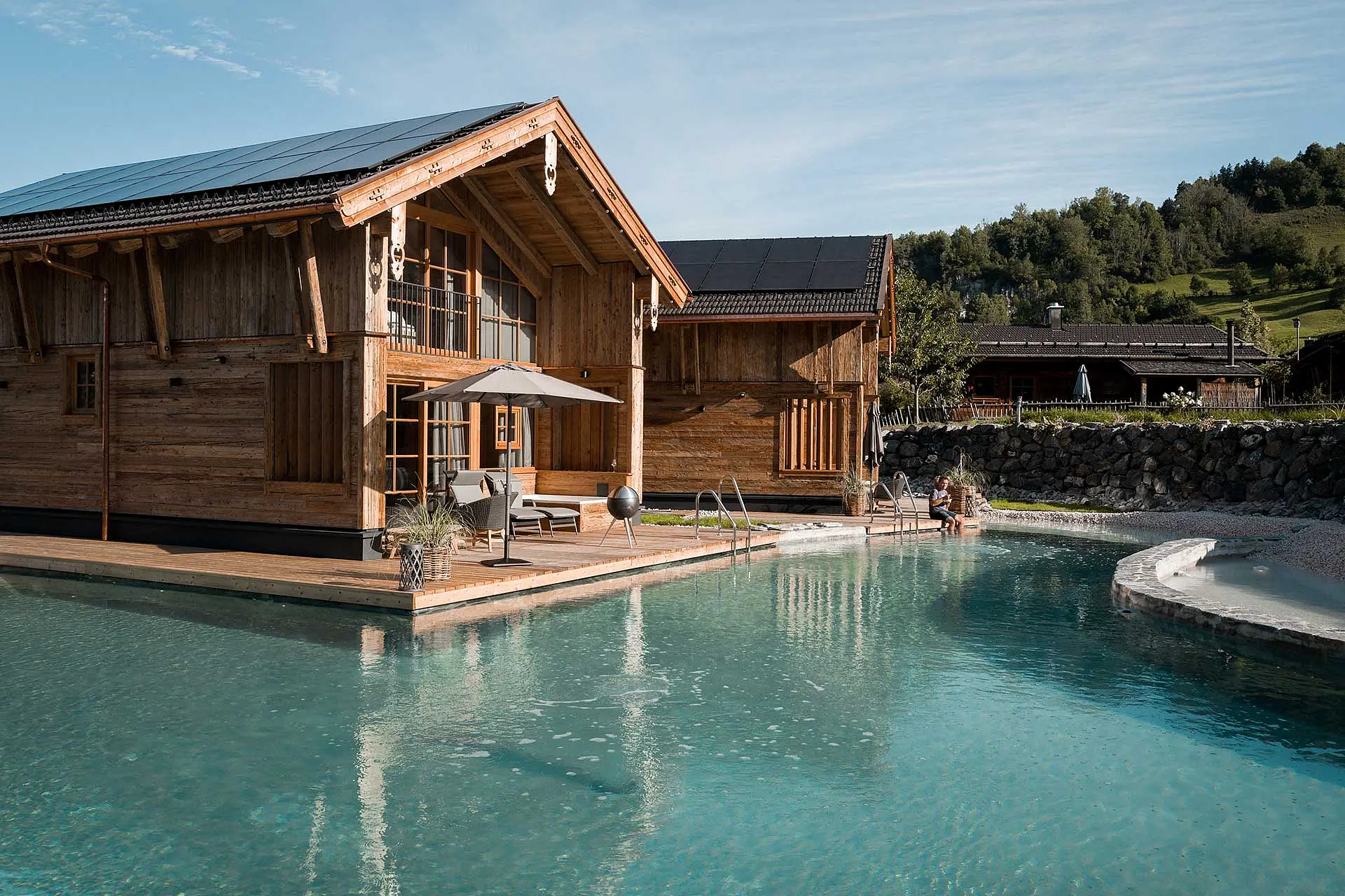 Ein modernes Holzhaus steht am Ufer eines glasklaren Pools, umgeben von grüner Landschaft. Eine Liege und ein Sonnenschirm sorgen für Entspannung, während eine Person im Wasser sitzt. Die Architektur verbindet Natur und zeitgemäßes Design.