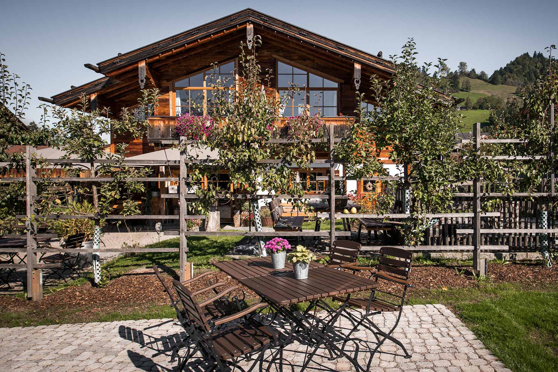 Ein rustikales Holzhaus mit großen Fenstern steht im Hintergrund. Im Vordergrund sind Tische und Stühle auf einem gepflasterten Bereich, umgeben von Pflanzen und Bäumen. Der Garten lädt zum Verweilen ein.