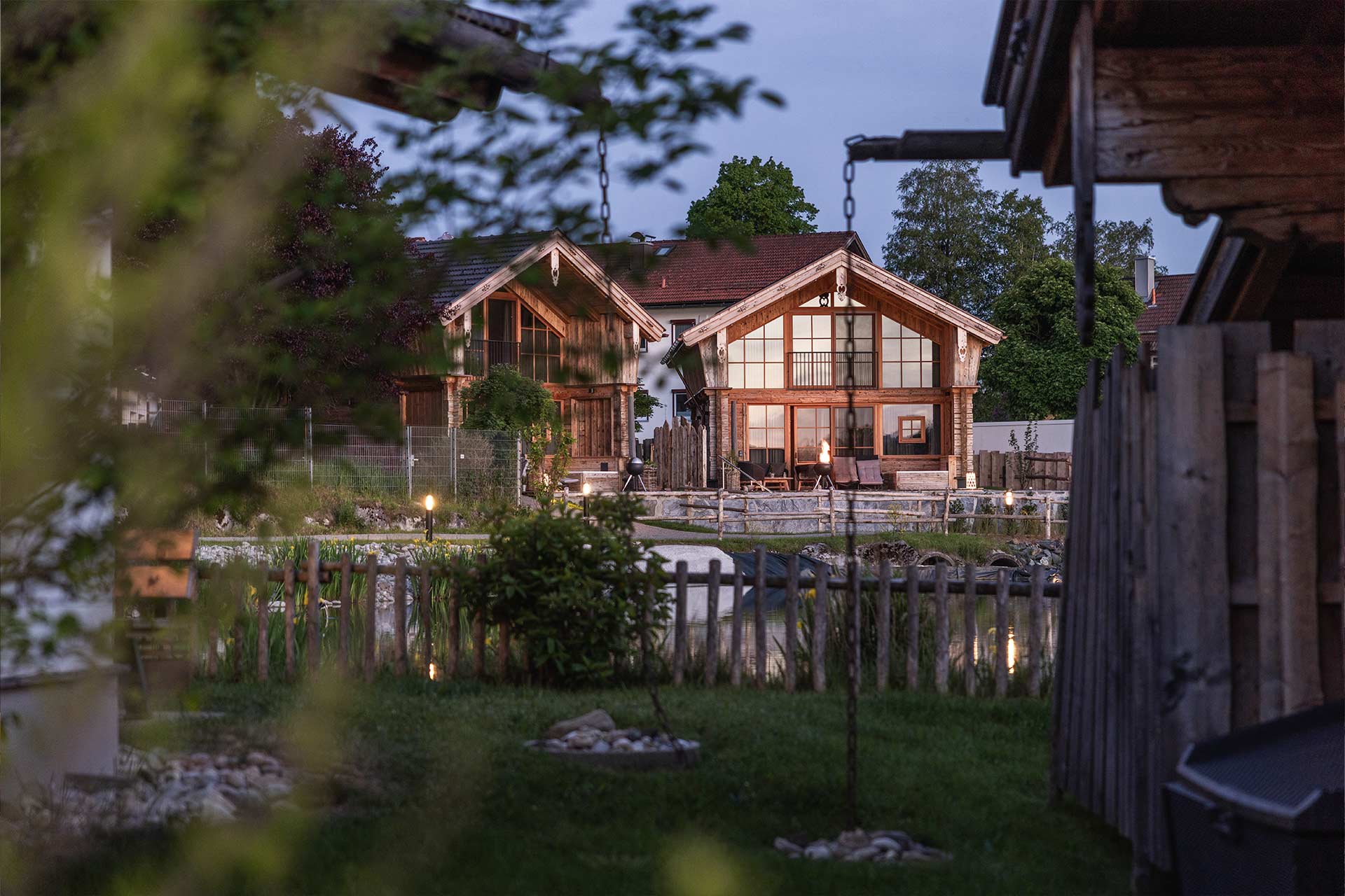 Zwei moderne Holzferienhäuser in einer ruhigen Umgebung, umgeben von einem Garten mit Büschen und Steinen. Im Hintergrund sieht man einen Teich oder Bach. Die Häuser haben große Fenster, die sanftes Licht ausstrahlen, während es dämmerig ist.