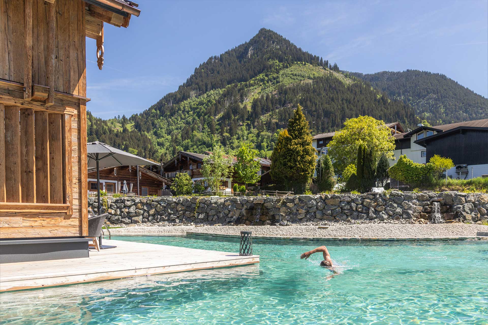 Ein Schwimmer taucht in einem klaren Pool vor einer malerischen Berglandschaft ein. Im Hintergrund sind grüne Hügel und Holzchalets zu sehen, während die Sonne am blauen Himmel scheint.