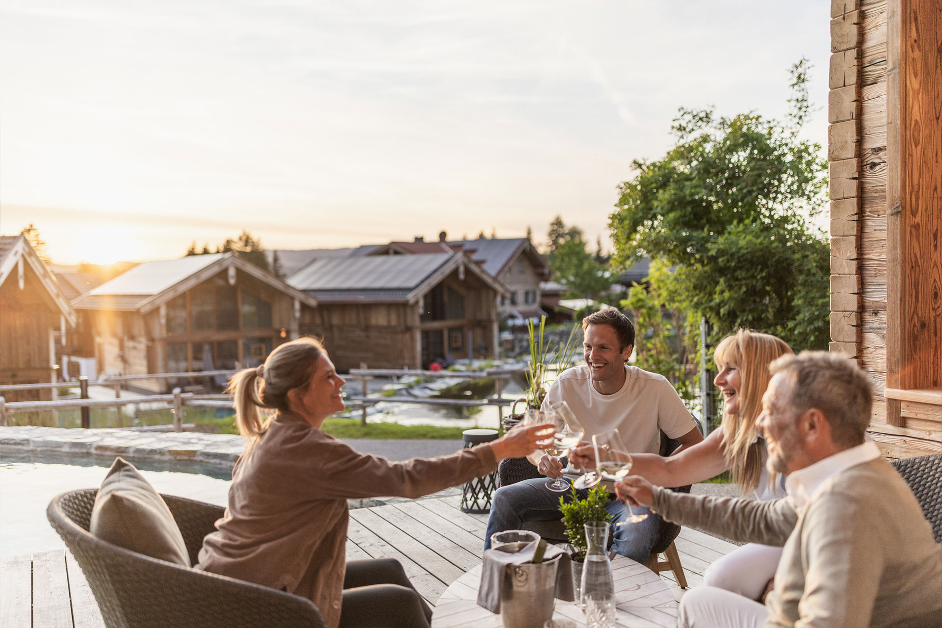 Eine Gruppe von vier Personen sitzt an einem Tisch auf einer Terrasse, umgeben von Natur. Sie stoßen mit Gläsern an und haben ein fröhliches Lächeln. Im Hintergrund sind Holzgebäude und ein Sonnenuntergang sichtbar.