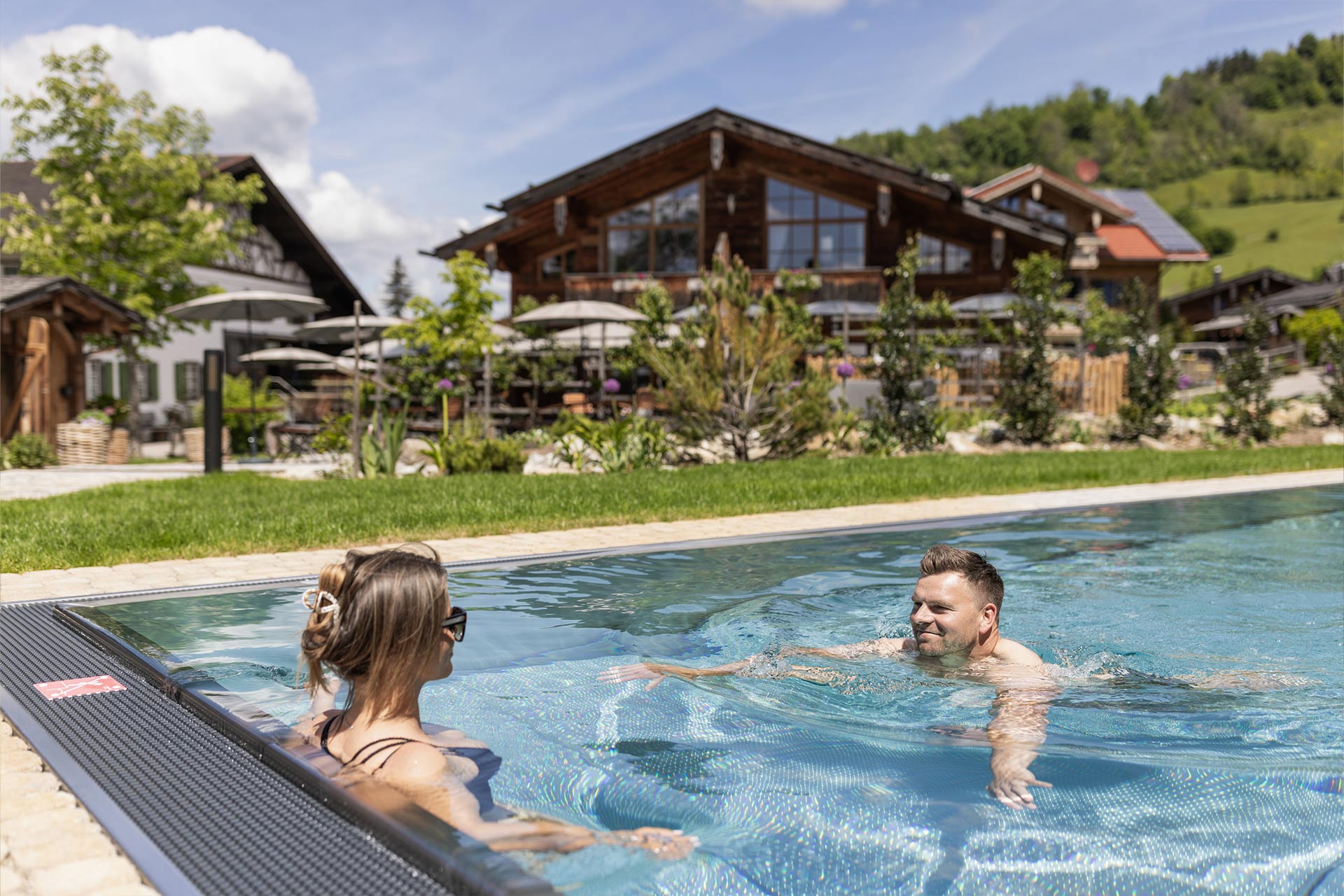 Ein Paar entspannt im Schwimmbad. Im Hintergrund sind ein rustikales Holzhaus und eine grüne Landschaft mit Bäumen zu sehen. Der Himmel ist blau mit einigen Wolken. Die Atmosphäre wirkt einladend und erholsam.