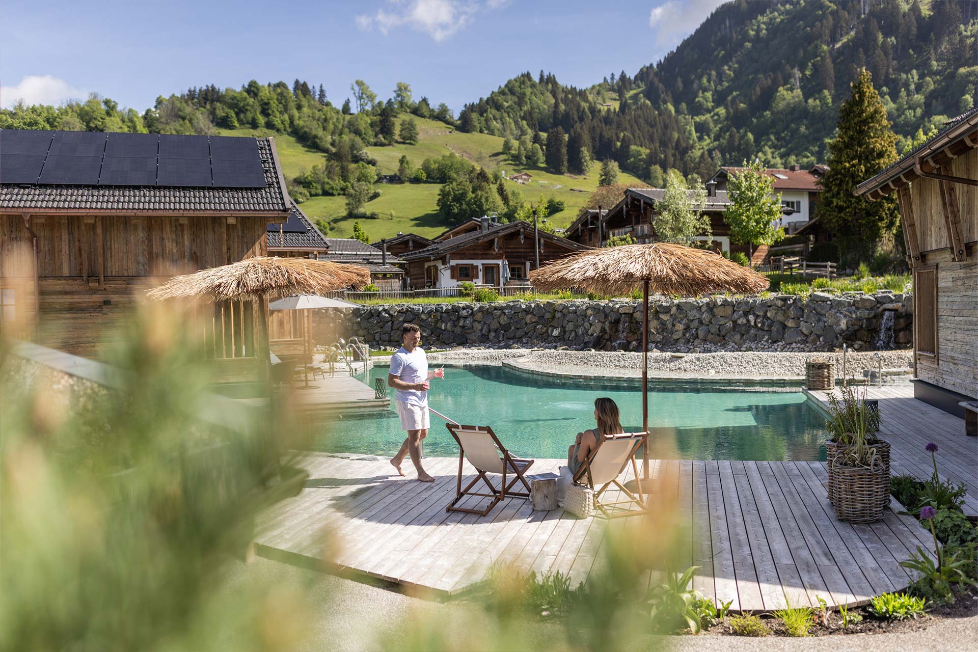 Eine idyllische Landschaft mit einem kleinen See und Holzterrassen, auf denen zwei Personen entspannen. Eine Person steht und hält ein Getränk, während die andere auf einer Liege sitzt. Im Hintergrund sind Grüne Hügel und verschiedene alpine Hütten sichtbar.