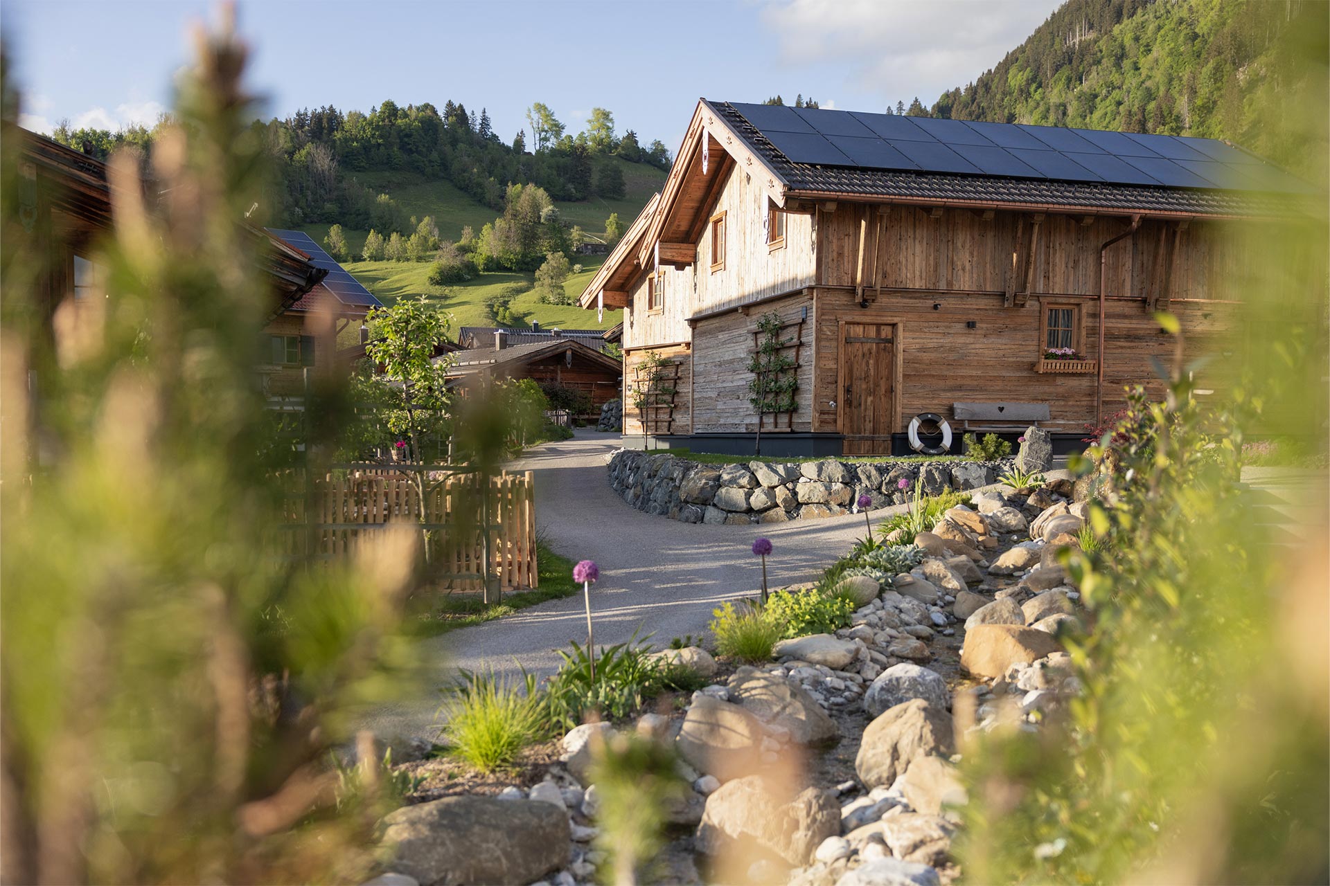 Eine malerische Landschaft mit Holzchalets umgeben von grünen Wiesen und Bergen. Im Vordergrund sind Steine und Pflanzen zu sehen, die einen Weg zu den Gebäuden führen. Die Gebäude haben Dächer mit Solarpanelen. Ideal für ländliche Erholung.