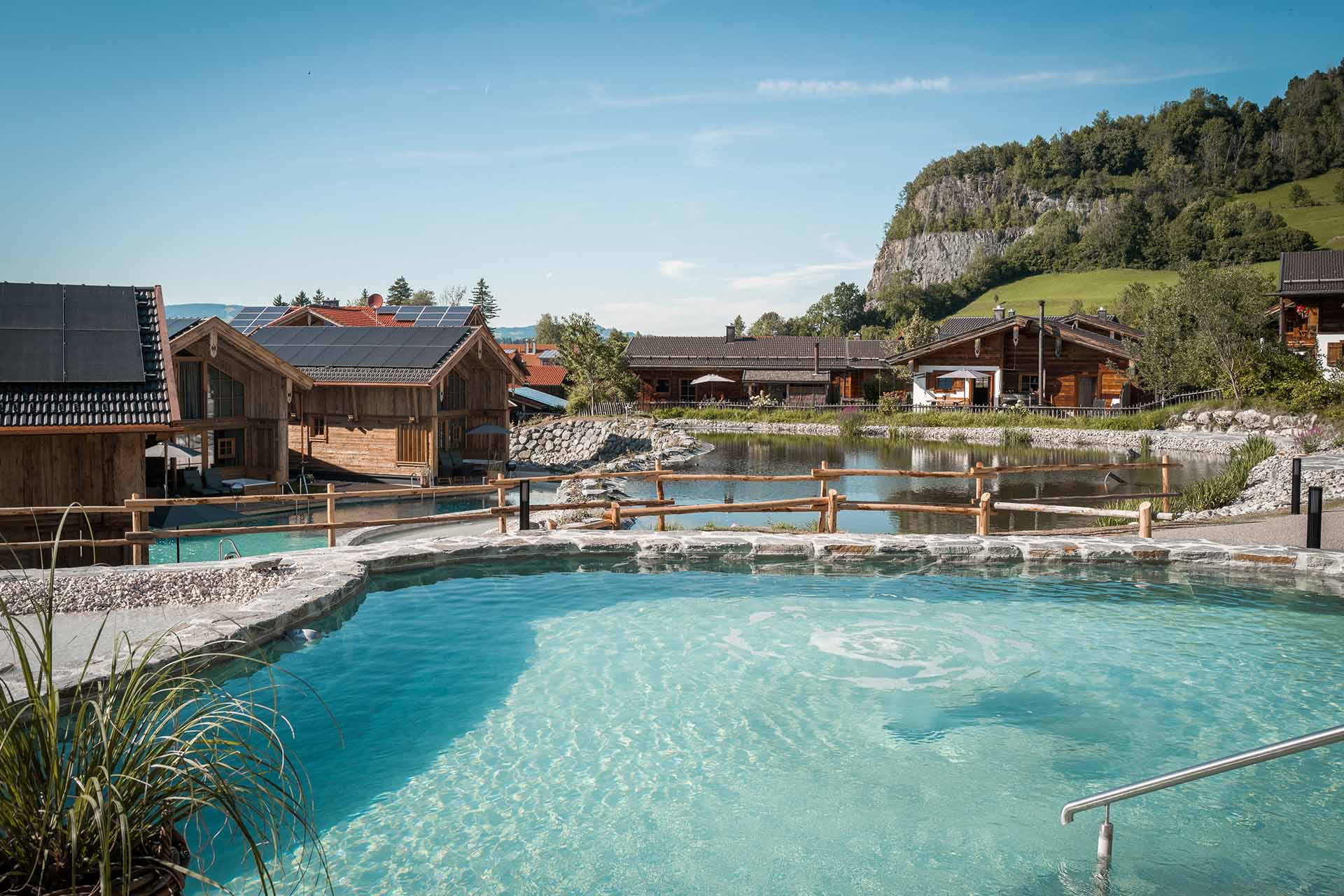 Ein idyllischer Außenbereich mit einem klaren, blauen Wasserbecken, umgeben von grünen Wiesen und Hügeln. Holzchalets stehen am Wasser, und die Landschaft strahlt eine ruhige, entspannende Atmosphäre aus.