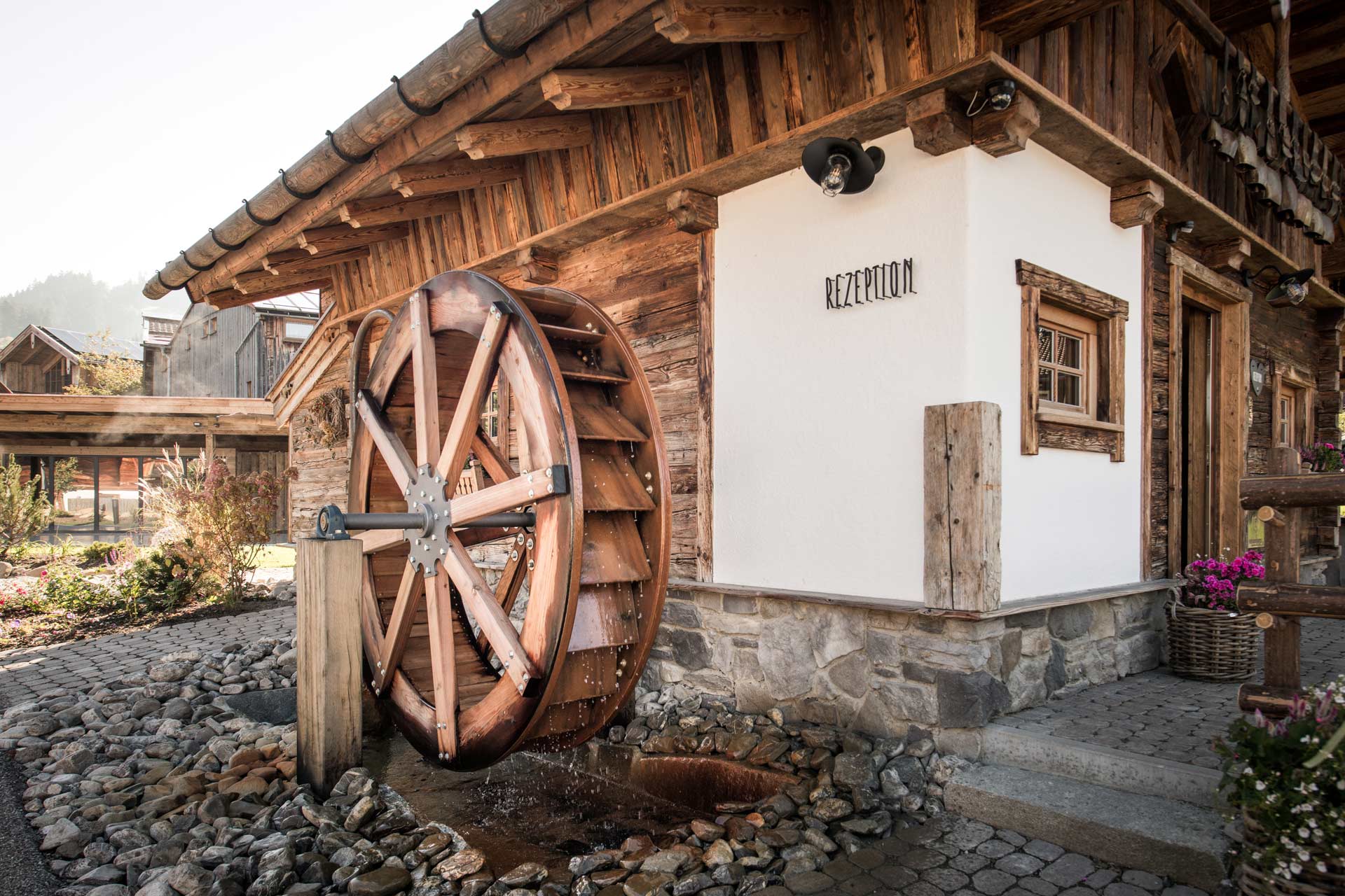 Ein traditionelles Holzrad mit Wasserantrieb steht neben einem rustikalen Empfangsgebäude. Die Fassade des Gebäudes ist aus Holz und Stein, mit einem Schild, das "Rezeption" anzeigt. Umgeben von Kies und Pflanzen, vermittelt die Szene eine ländliche, einladende Atmosphäre.