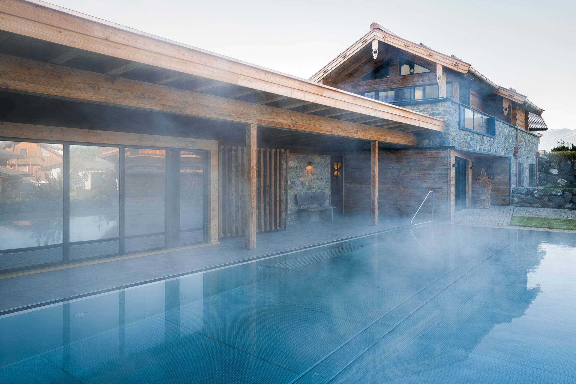 Ein moderner Infinity-Pool mit dampfender Wasseroberfläche, umgeben von einem Holz- und Steinbau im Hintergrund. Der Gebäudebereich hat große Fenster und ein Dach mit traditionellem Design. Die Atmosphäre ist ruhig und einladend.