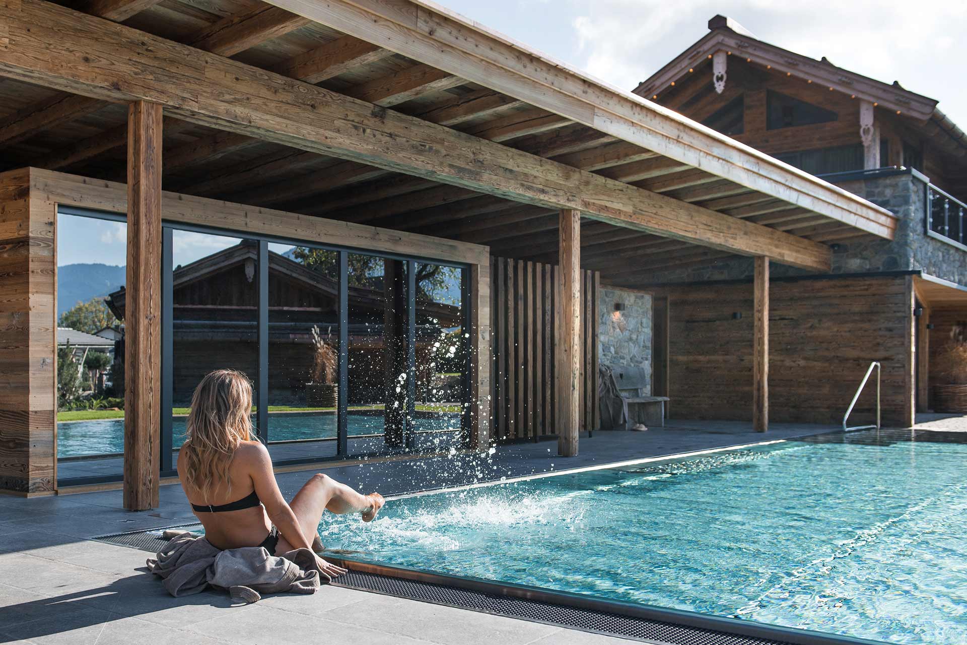 Eine Frau sitzt am Rand eines Pools, zieht ihr Bein ins Wasser und spritzt. Der Bereich ist von holzverkleideten Wänden umgeben und hat große Fenster, die einen Blick auf die Natur bieten. Sonnenlicht fällt auf die Szenerie, die entspannt und einladend wirkt.