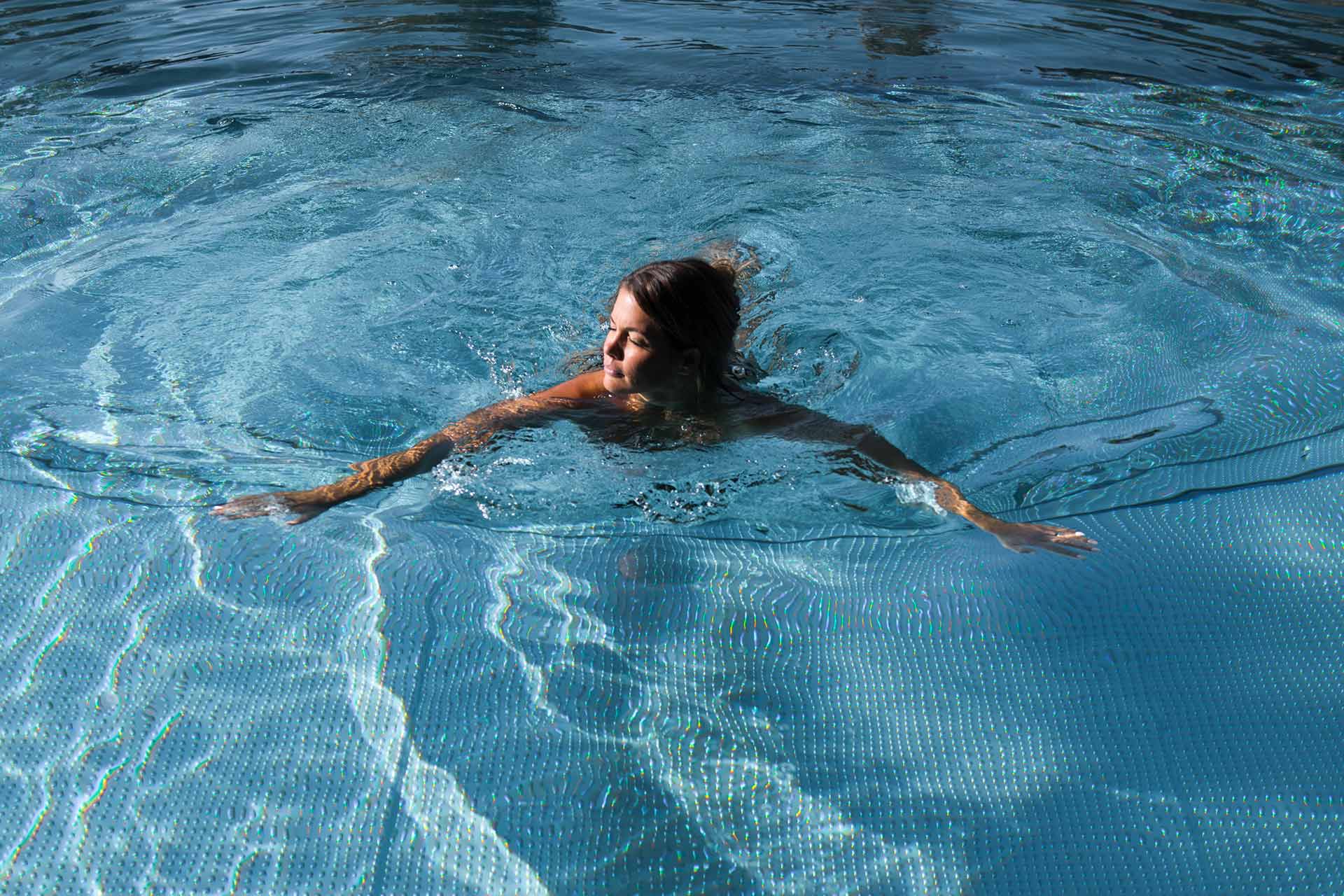 Eine Frau schwimmt entspannt in einem klaren Pool. Ihr Haar ist nass, und sie bewegt sich sanft im Wasser, wobei die Wellen um sie herum tanzen. Die Szenerie vermittelt ein Gefühl von Ruhe und Erholung.