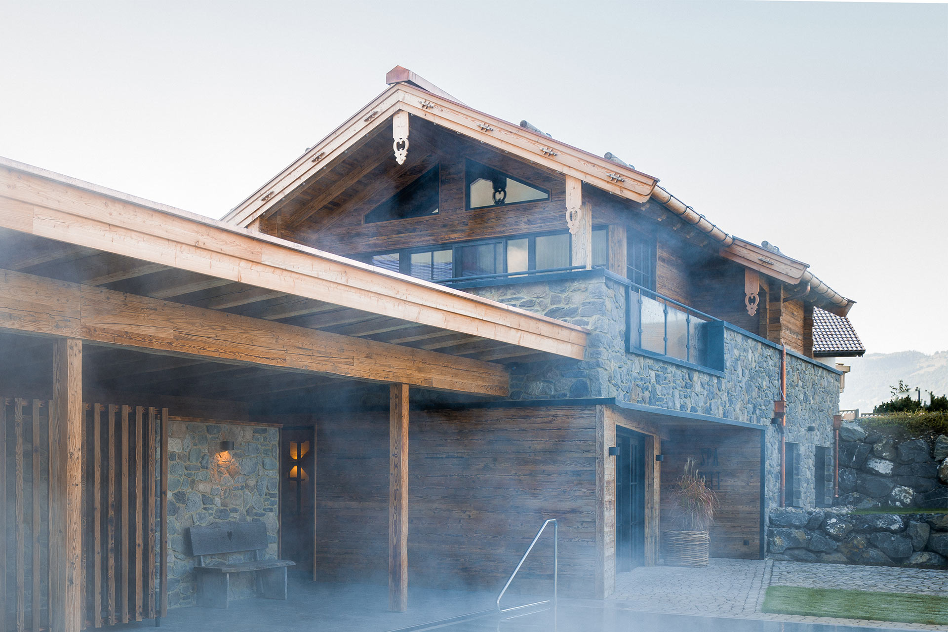 Ein modernes Haus mit einem Holz- und Steinmix, das auf einem Grundstück in einer bergigen Umgebung steht. Die Fassade zeigt große Fenster und ein Dach mit Holzverzierungen. Leichter Nebel umgibt das Gebäude und verleiht der Szene eine ruhige, stimmungsvolle Atmosphäre.