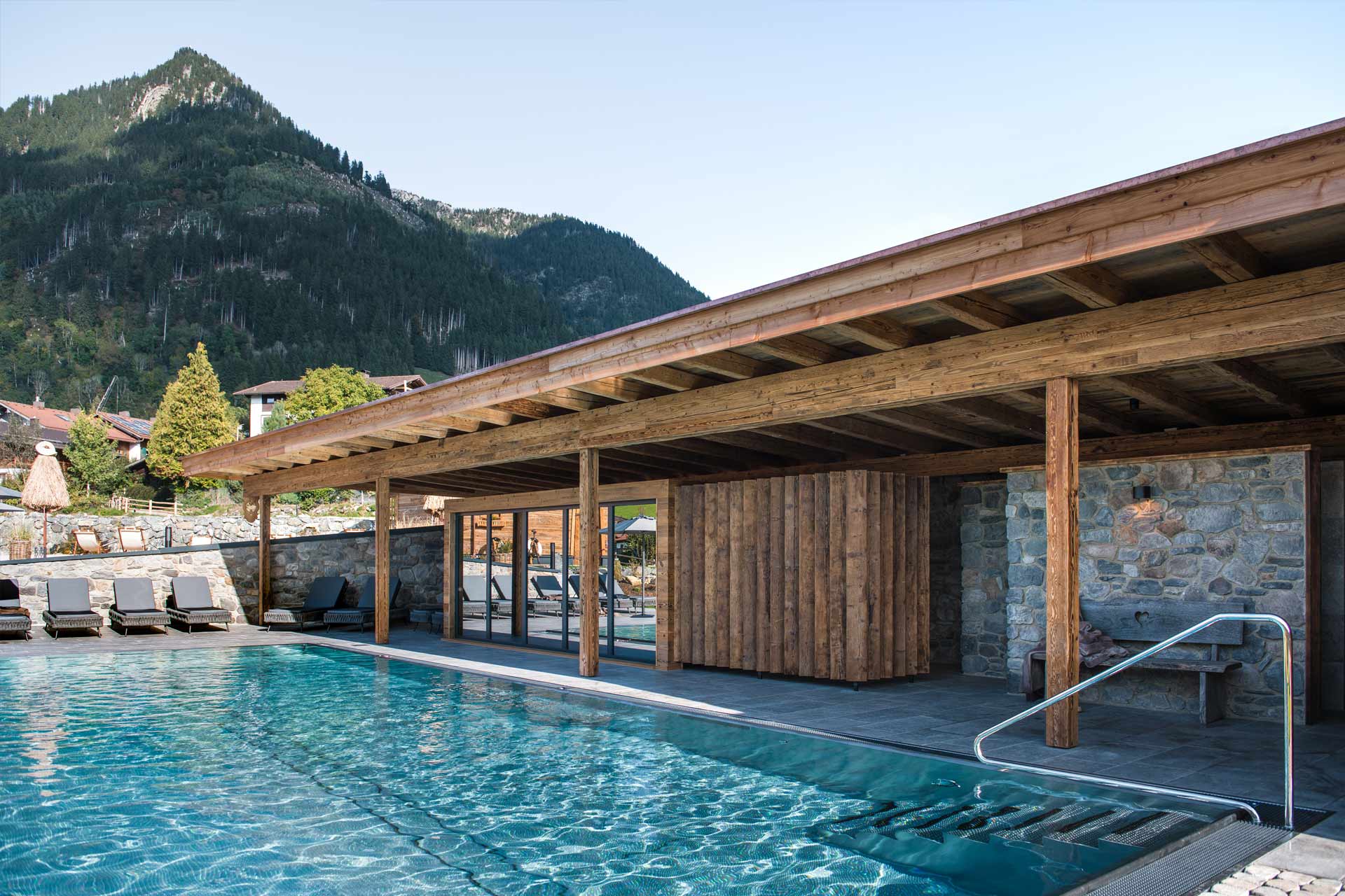 Ein modernes Schwimmbad mit glattem Wasser, umgeben von Holz- und Steinarchitektur. Im Hintergrund sind bewaldete Berge sichtbar. Auf der rechten Seite befindet sich ein Zugang zum Wasser mit einem Handlauf. Der Bereich wirkt einladend und entspannt.