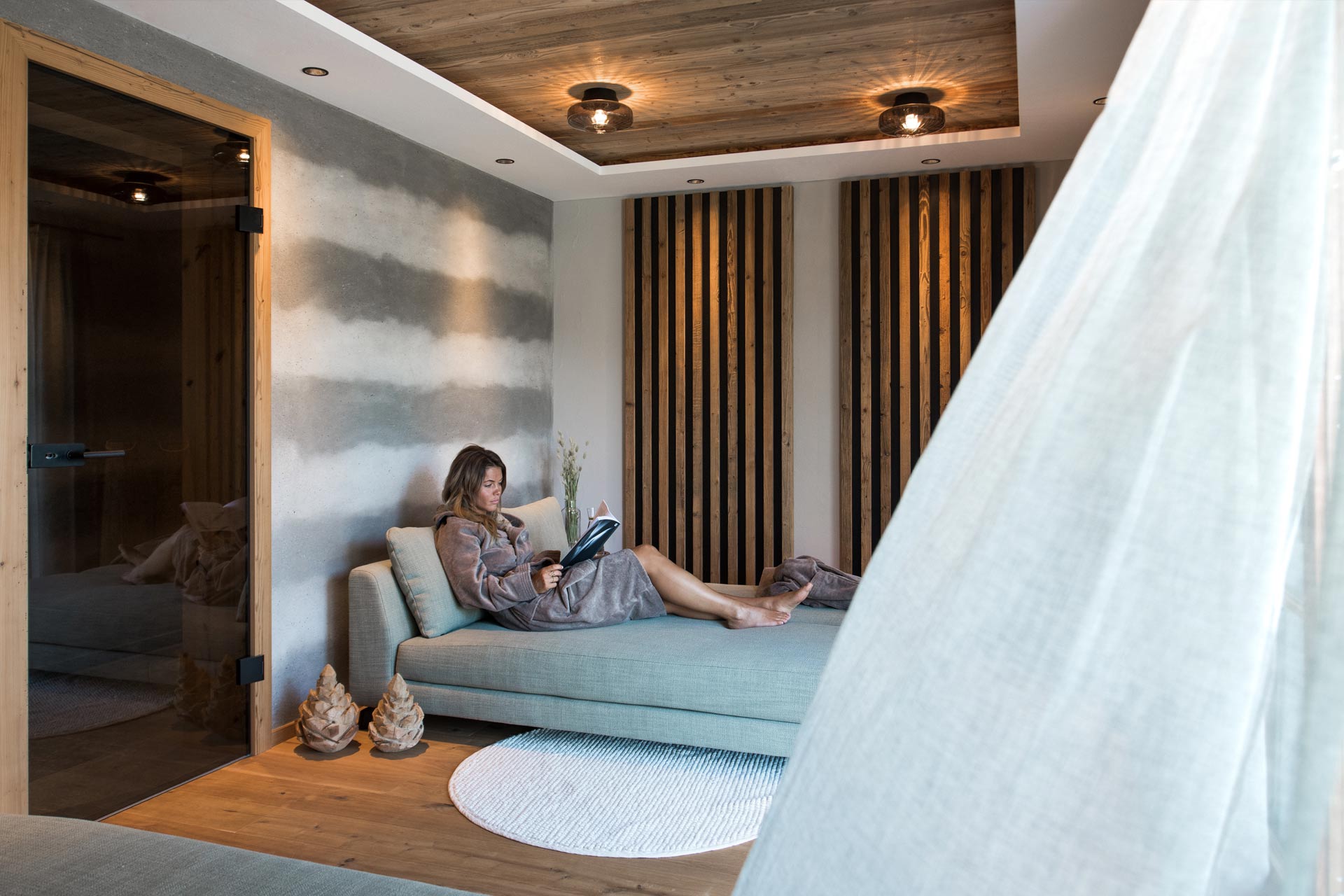 Eine Frau sitzt entspannt auf einer Couch in einem modernen Raum. Sie trägt einen warmen Bademantel und hält ein Buch. Der Raum ist mit Holzverkleidungen und sanfter Beleuchtung gestaltet, was eine ruhige Atmosphäre schafft. Ein runder Teppich liegt auf dem Holzfußboden.