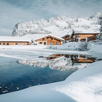 Eine malerische Winterlandschaft mit verschneiten Holzhäusern, umgeben von schneebedeckten Hügeln. Ein ruhiger Teich spiegelt die Umgebung wider und verstärkt die friedliche Atmosphäre der Szene.