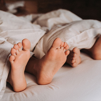 Die Abbildung zeigt zwei Paar nackte Füße, die aus einer Decke herausschauen. Die Füße sind auf einem Bett positioniert und vermitteln ein Gefühl von Entspannung und Nähe.