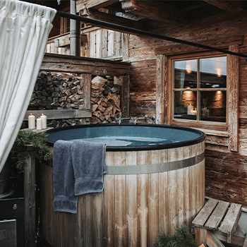 A cozy wooden hot tub with a dark blue interior is situated in a rustic outdoor setting. It features a light gray towel draped over the edge and is surrounded by wooden walls and stacks of firewood. Soft candlelight glows from a nearby window.