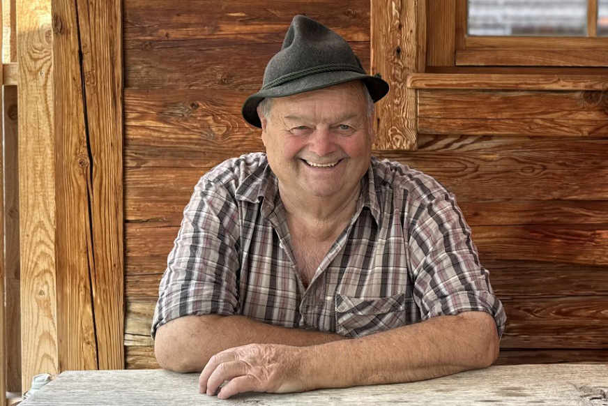 Ein älterer Mann mit einem grünen Hut sitzt an einem Holztisch. Er trägt ein kariertes Hemd und lächelt freundlich. Im Hintergrund sind Holzwände sichtbar, die eine rustikale Atmosphäre schaffen.