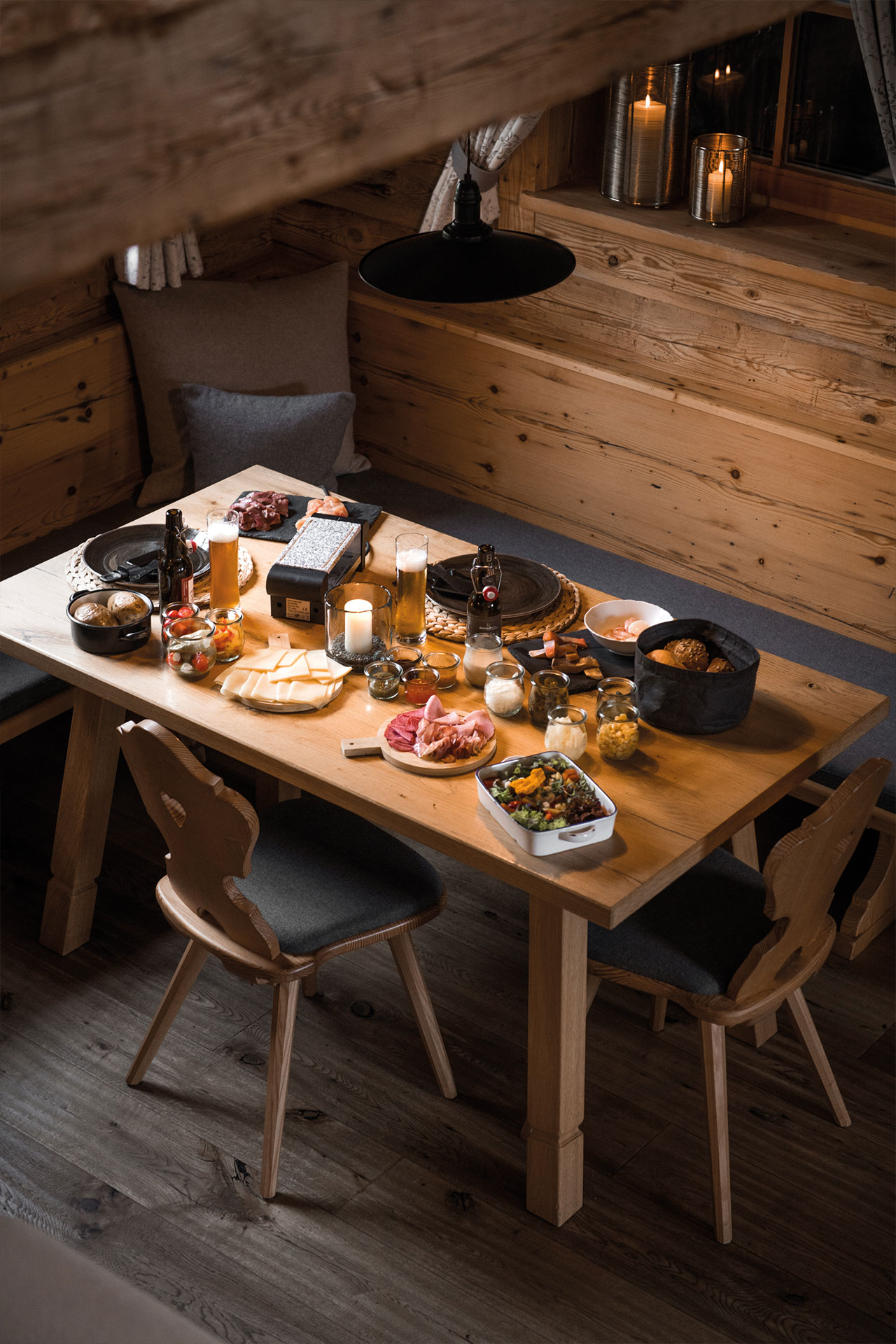 A rustic wooden table is set for a meal, featuring various dishes including salads, meats, and bread. Candles provide soft lighting in a cozy, warmly decorated space, enhancing the inviting atmosphere of the dining area.