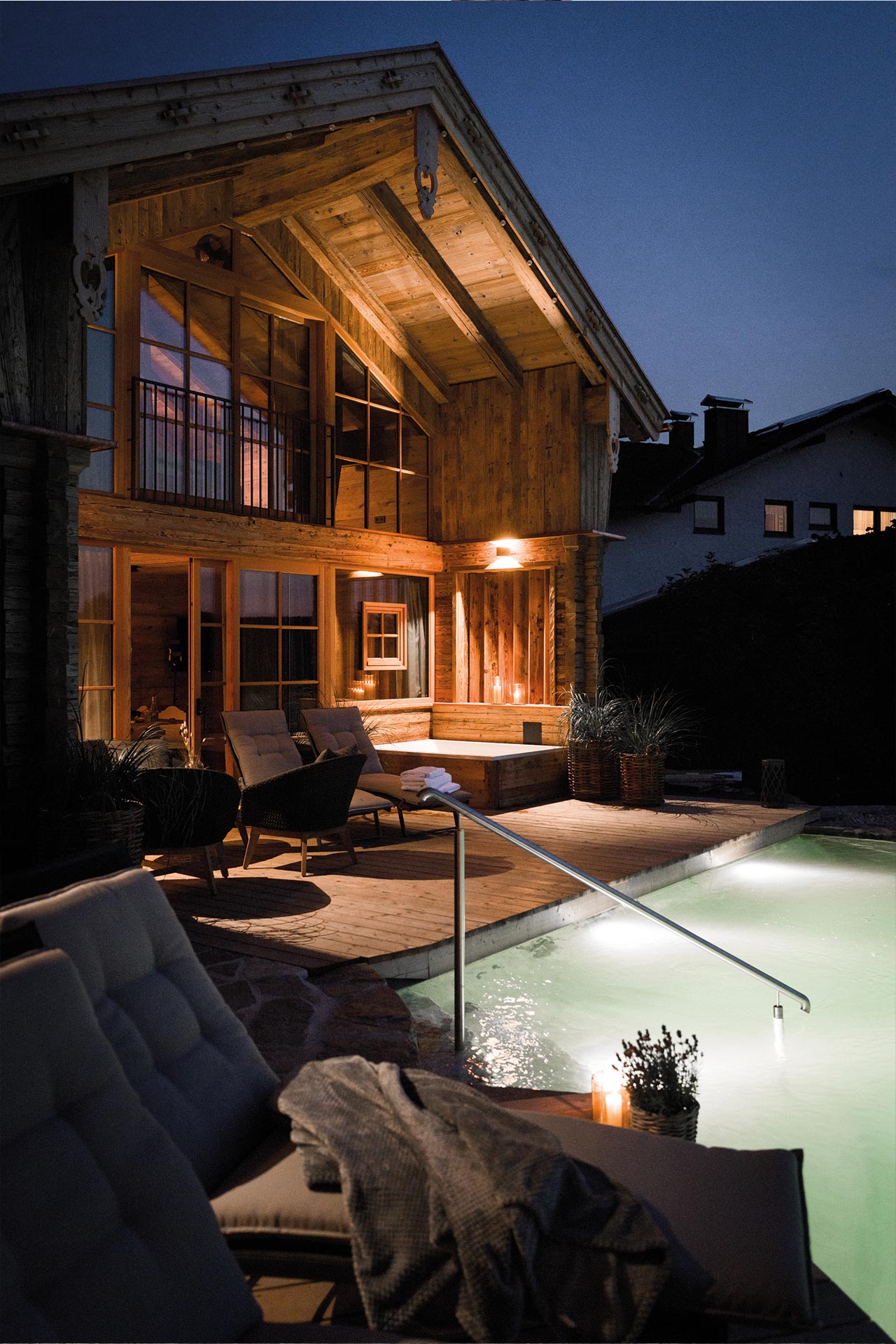 Ein modern gestaltetes Holzhaus bei Nacht, beleuchtet von warmen Lichtern. Im Vordergrund befindet sich ein Swimmingpool, daneben bequeme Sitzmöbel und eine gemütliche Decke. Die Atmosphäre ist ruhig und einladend, mit Pflanzen, die den Außenbereich ergänzen.